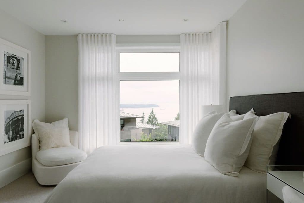 The Peak at Mulgrave Park – Bright bedroom with an all-white bed, framed black-and-white photos on the wall, an armchair, and a large window looking out at rooftops and the ocean.