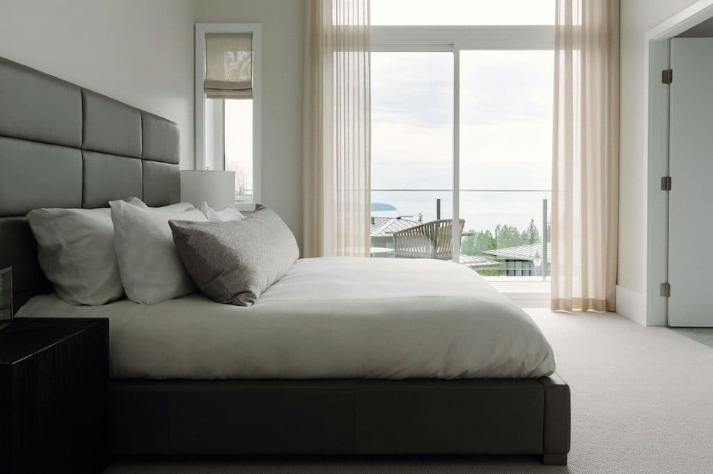The Peak at Mulgrave Park – Bedroom with a dark leather headboard, layered white and grey bedding, and floor-to-ceiling sliding glass doors opening to a balcony with an ocean view.
