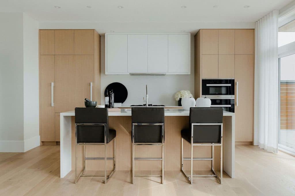 The Peak at Mulgrave Park – Light wood kitchen with white upper cabinets, a large island with three black bar stools, and a built-in oven, opening to a balcony.