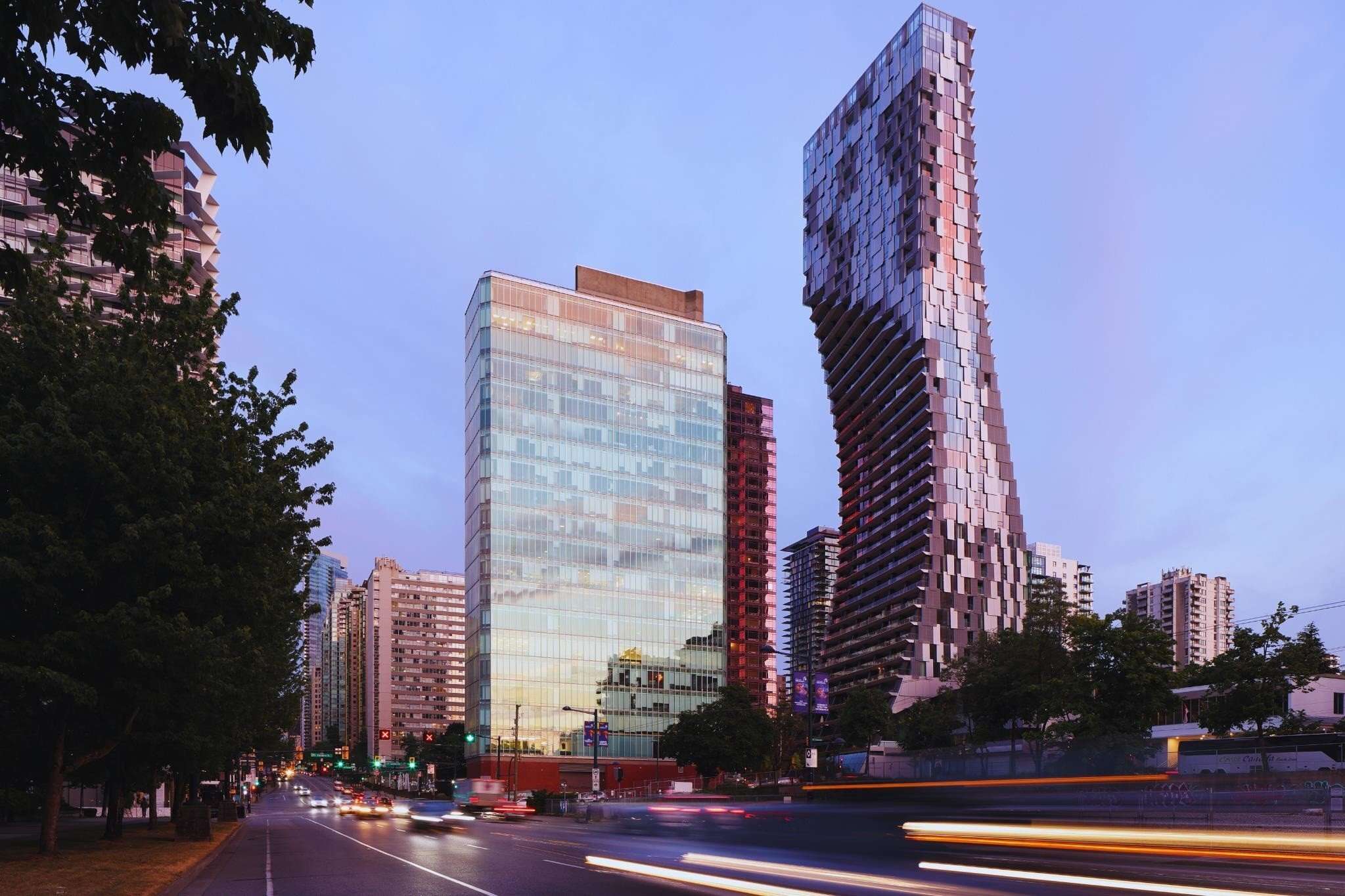 Alberni by Kengo Kuma – A ground-level street view at twilight featuring a striking, twisting glass residential high-rise alongside a modern glass office building. Vehicle light trails streak across the city roadway in the foreground, framing the architectural towers against a soft dusk sky.