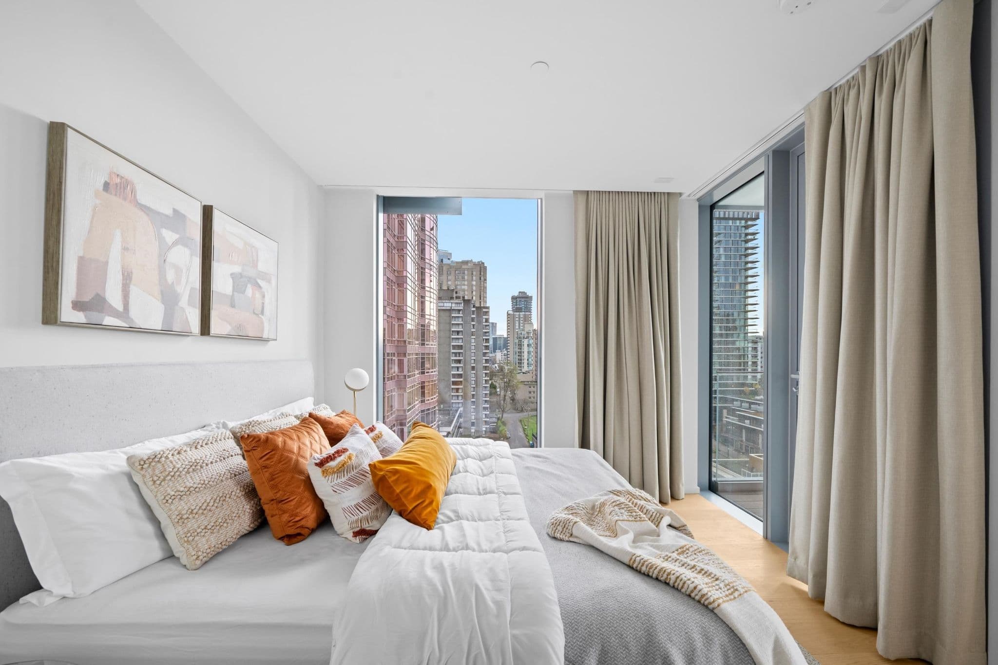 Alberni by Kengo Kuma – A modern bedroom features a neatly made bed styled with white linens and warm orange accent pillows against a light gray upholstered headboard. Large floor-to-ceiling windows and a sliding glass door let in bright daylight, offering views of surrounding high-rise city buildings.