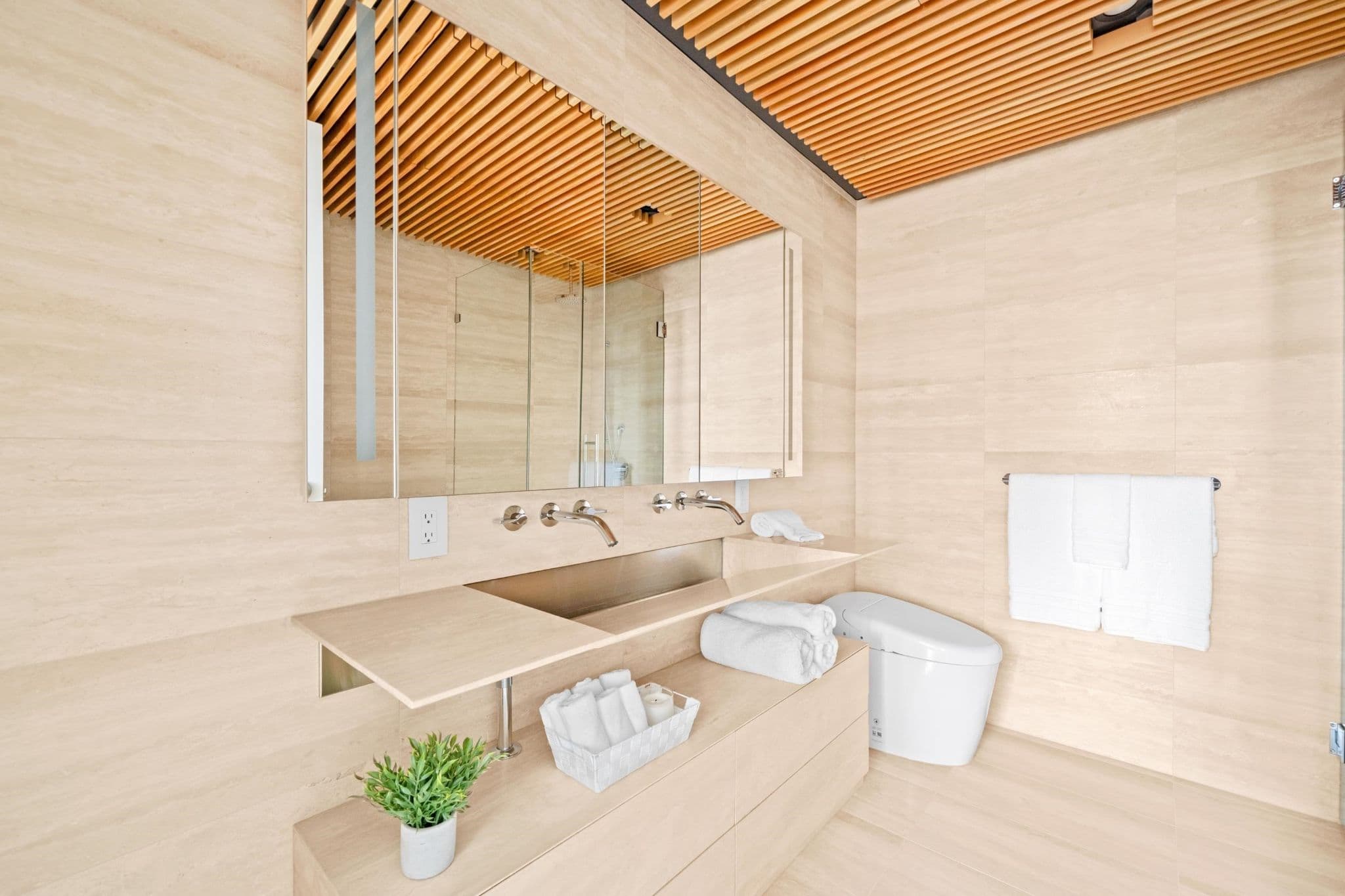 Alberni by Kengo Kuma – A modern bathroom featuring beige travertine-style wall and floor tiles paired with a warm wood slatted ceiling. It includes a floating vanity with a integrated trough sink, wall-mounted silver faucets, a large mirror, and a sleek white bidet toilet.