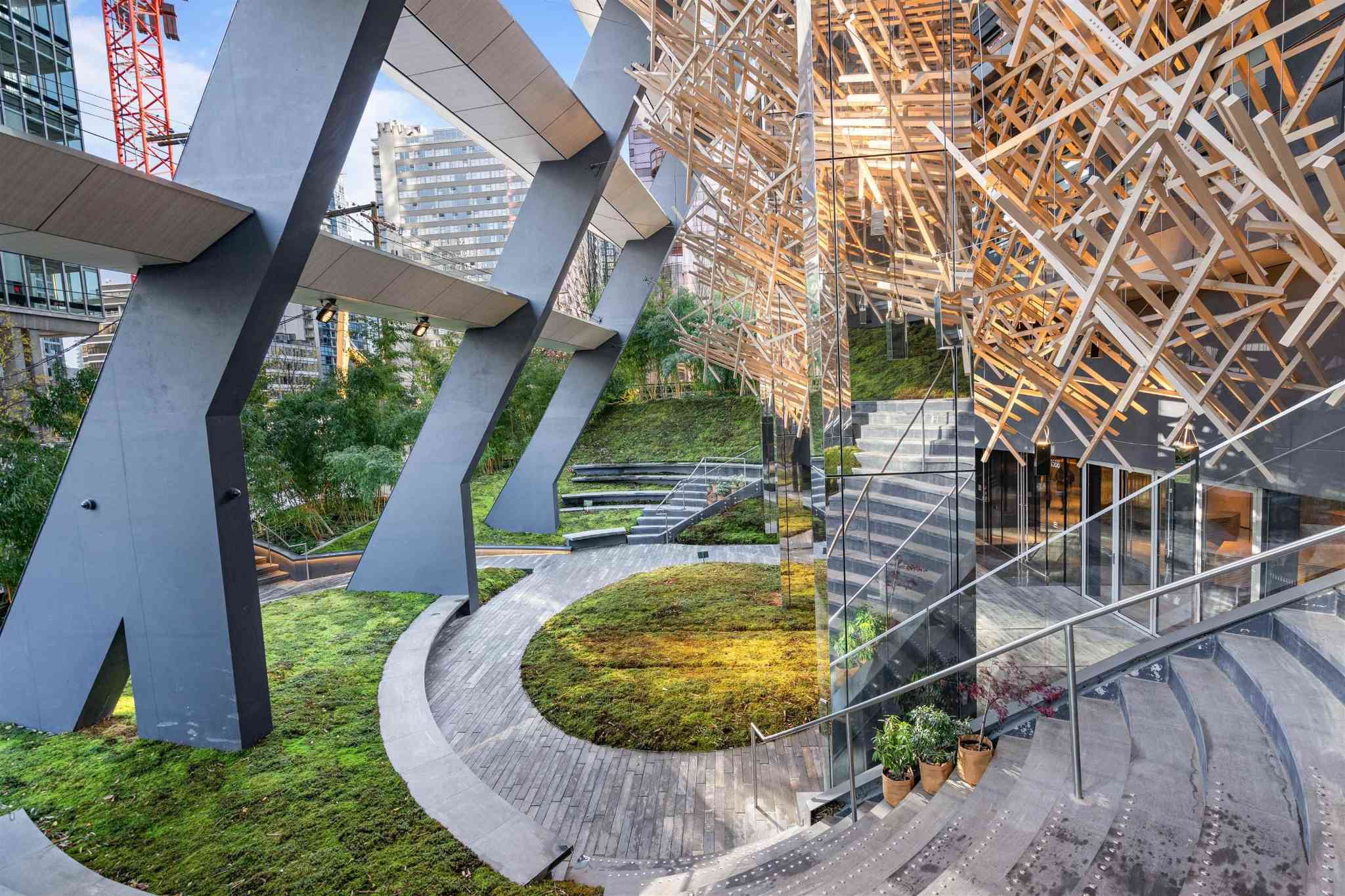 Alberni by Kengo Kuma – An architectural outdoor plaza features angled dark steel support columns, a grassy moss-covered terraced lawn, and curved stone pathways. A striking geometric lattice of light wood beams suspends over reflective glass walls and concrete staircases, setting an artistic, modern tone under daytime light.