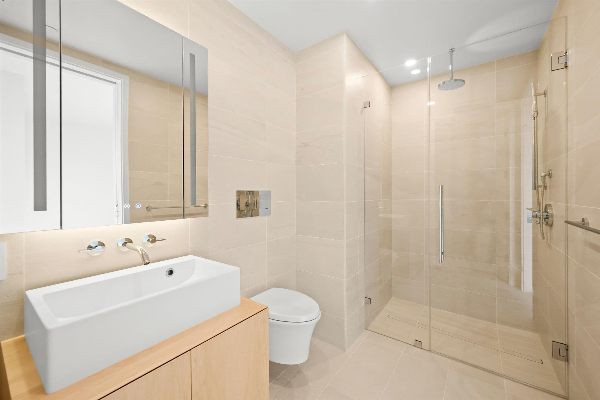 Alberni by Kengo Kuma – A modern, minimalist bathroom features warm beige stone wall tiles, a light wood vanity, and a rectangular vessel sink with wall-mounted chrome fixtures. Beside a wall-hung toilet, a seamless glass-enclosed walk-in shower includes a rain showerhead and handheld attachment.