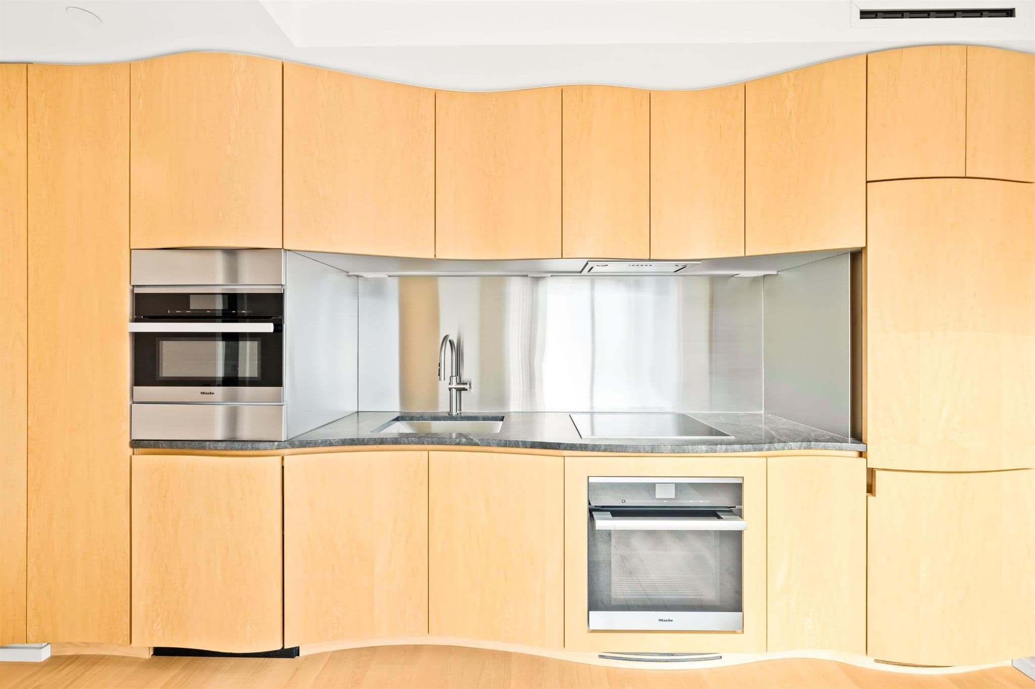 Alberni by Kengo Kuma – A modern residential kitchen features custom light wood cabinetry with a distinct gently undulating, curved profile along the top and bottom edges. The cooking area is framed by a stainless steel backsplash and dark granite countertop, fully equipped with integrated stainless steel Miele appliances including a built-in oven, microwave, and electric cooktop.