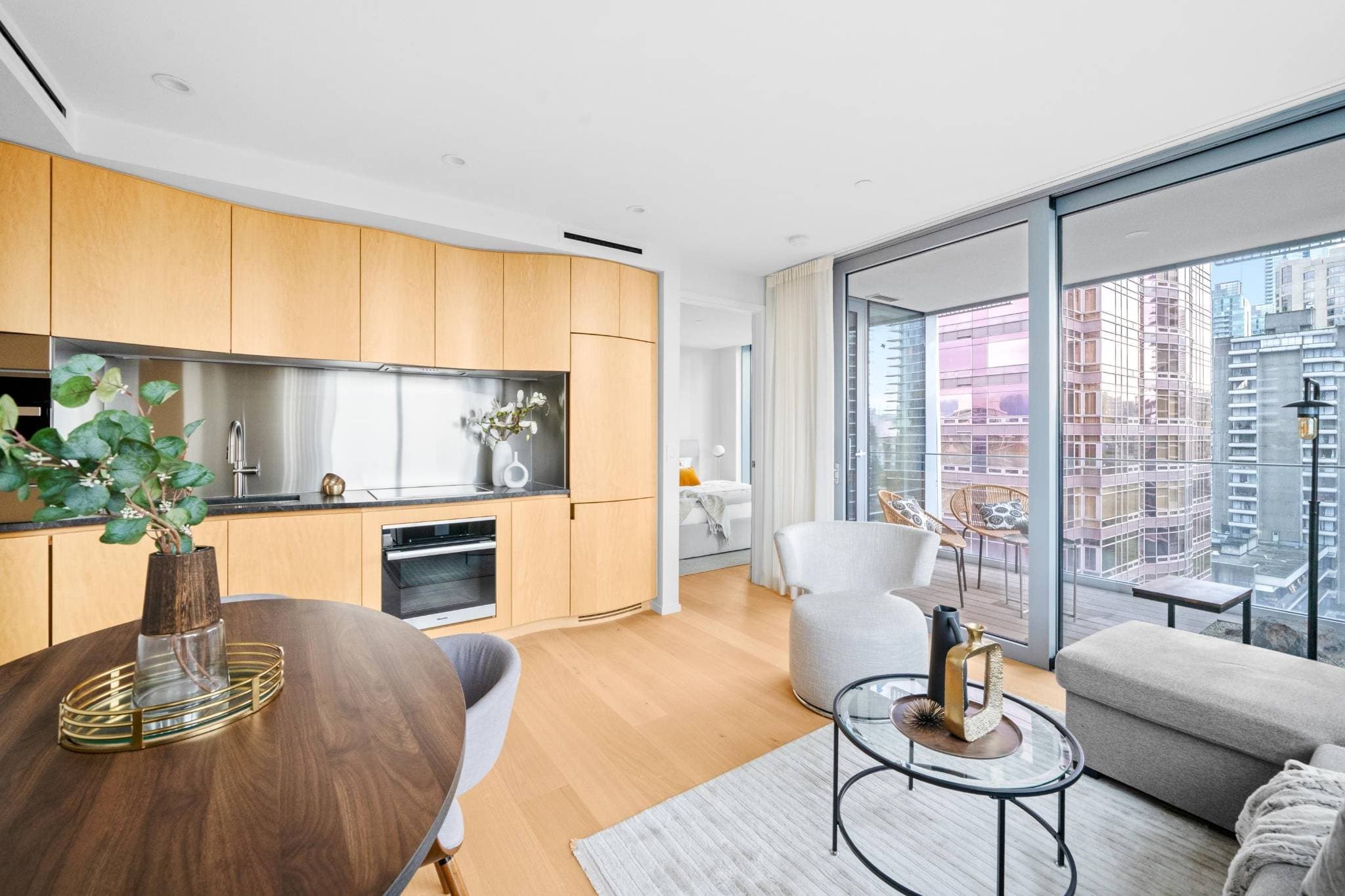 Alberni by Kengo Kuma – A modern open-concept condo interior featuring light wood kitchen cabinetry with integrated appliances, a small dining area, and a bright living space. Large floor-to-ceiling glass sliding doors lead out to a private balcony overlooking neighbouring city high-rises.
