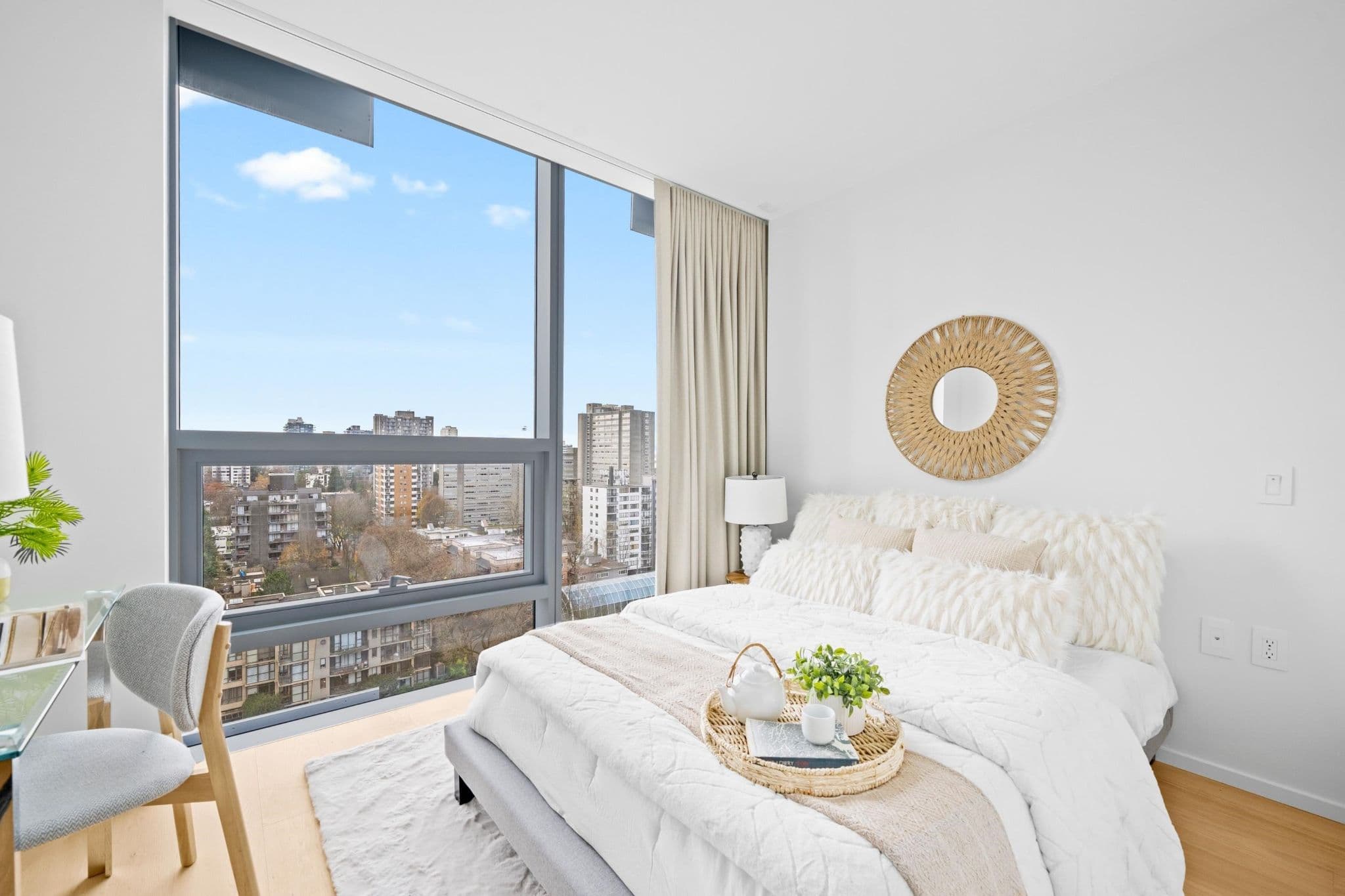 Alberni by Kengo Kuma – A bright, modern bedroom featuring a bed made with textured white bedding and plush pillows, positioned next to a large floor-to-ceiling window. The window offers an elevated view of surrounding city buildings and trees under a clear blue sky, while neutral curtains and a woven mirror complement the light wood flooring.