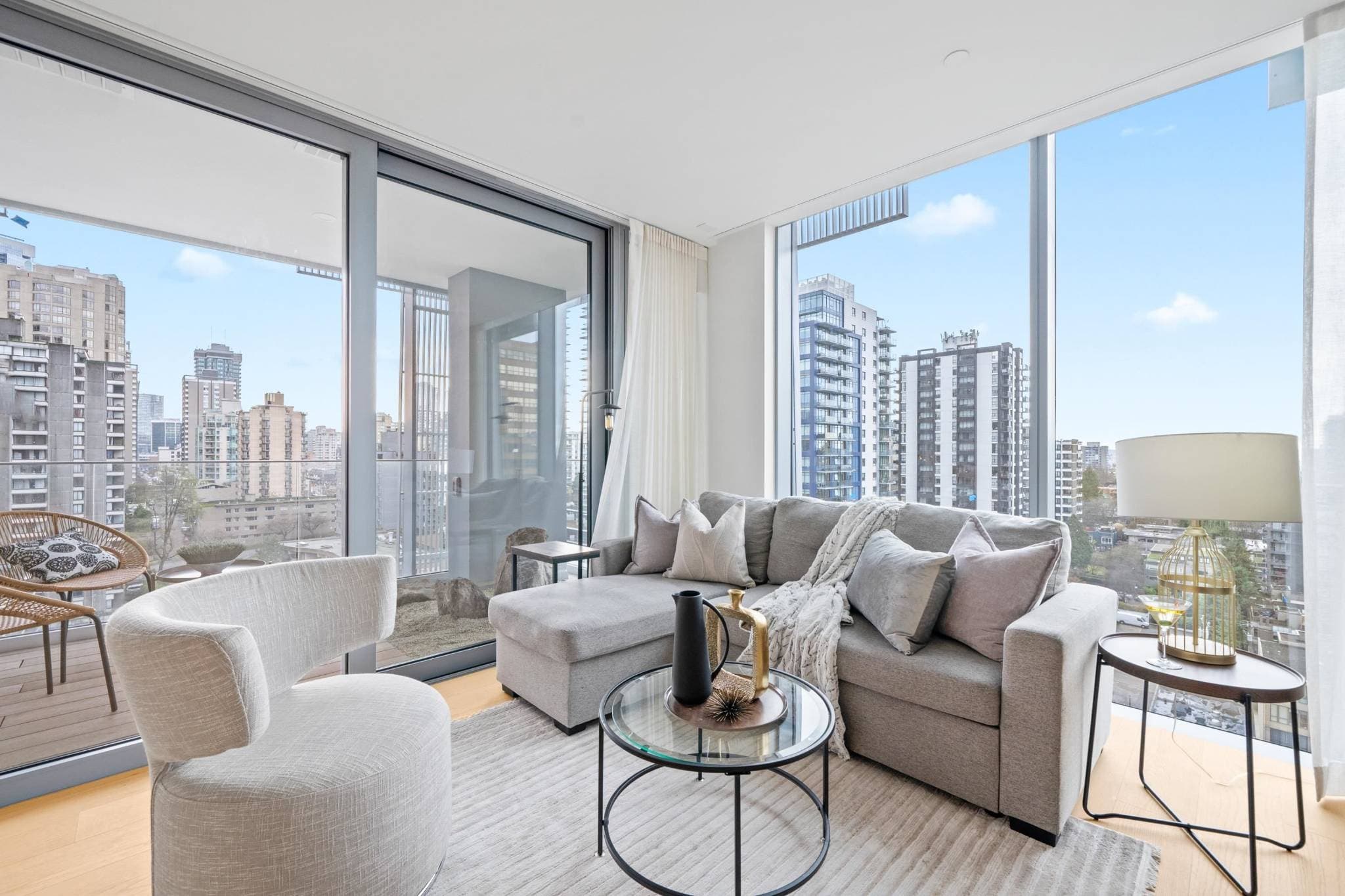 Alberni by Kengo Kuma – A modern corner living room features light grey seating, a textured area rug, and floor-to-ceiling windows that fill the space with daylight. Sliding glass doors lead out to a private balcony overlooking a dense cityscape under a clear blue sky.