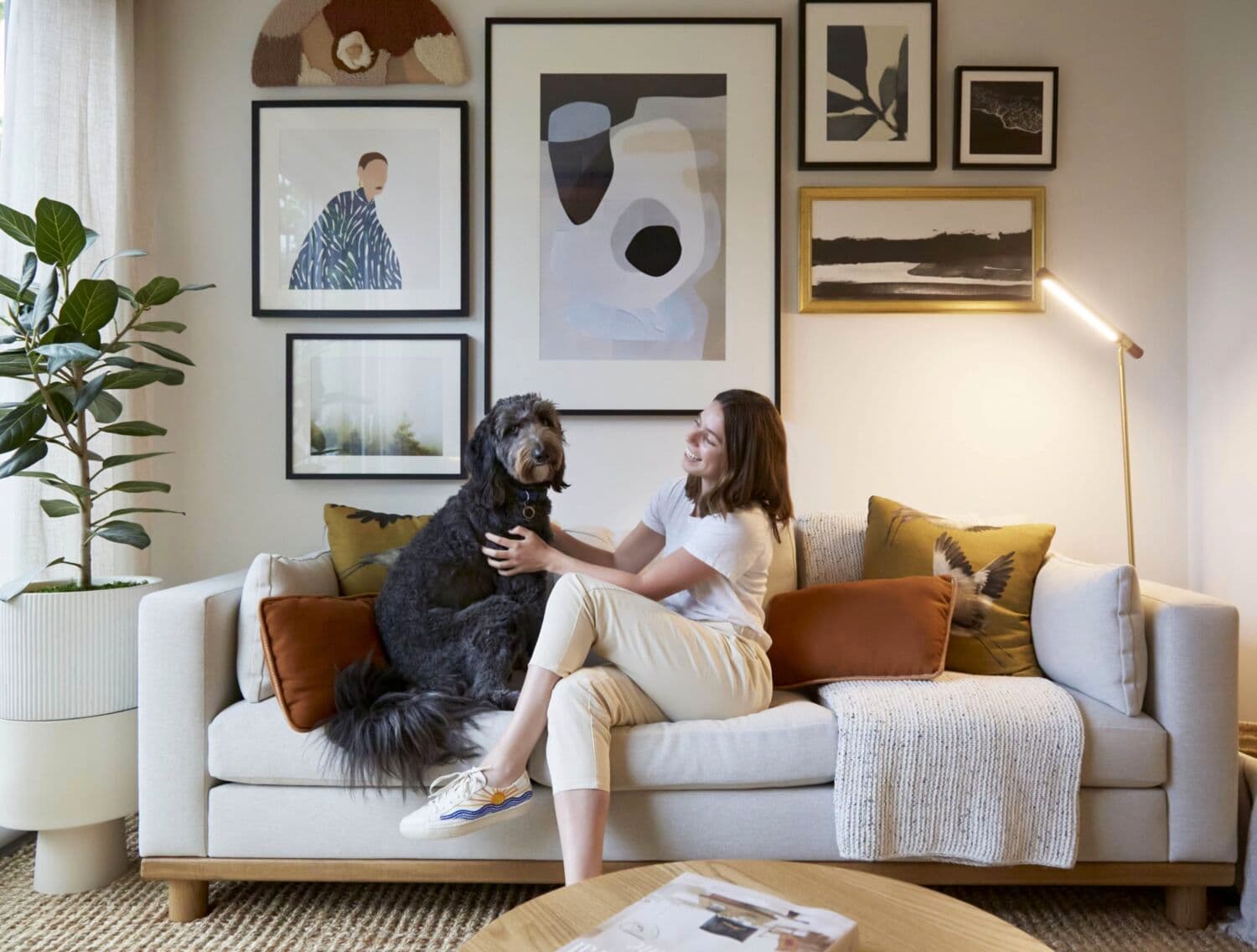 Moody Yards – A living room with a beige sofa, gallery wall of framed art, and a woman and her shaggy dog sitting together on the couch.