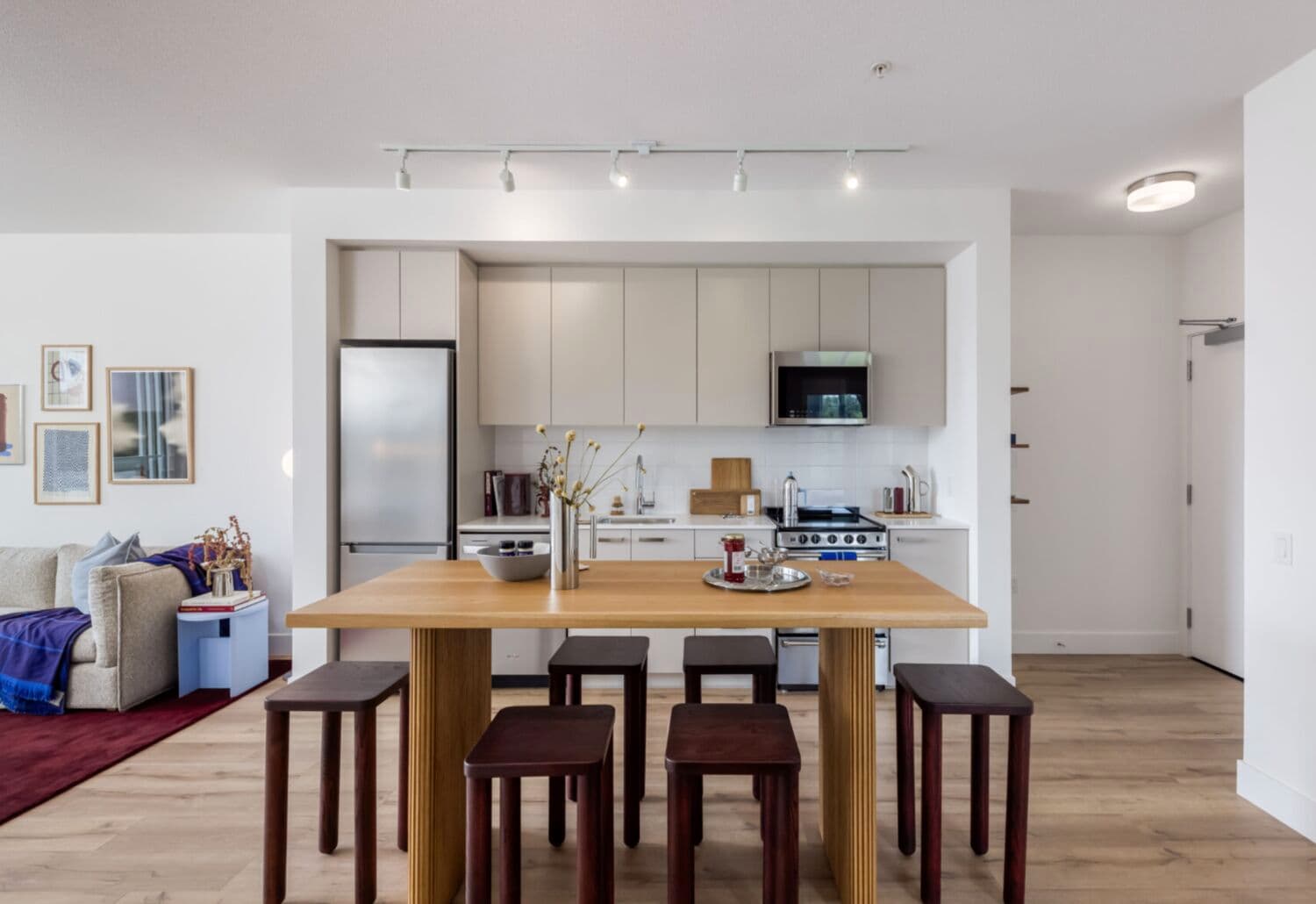 Moody Yards – A kitchen with light grey cabinetry, a stainless fridge and range, and a wood dining table with dark stools, opening to a living room and front door beyond.