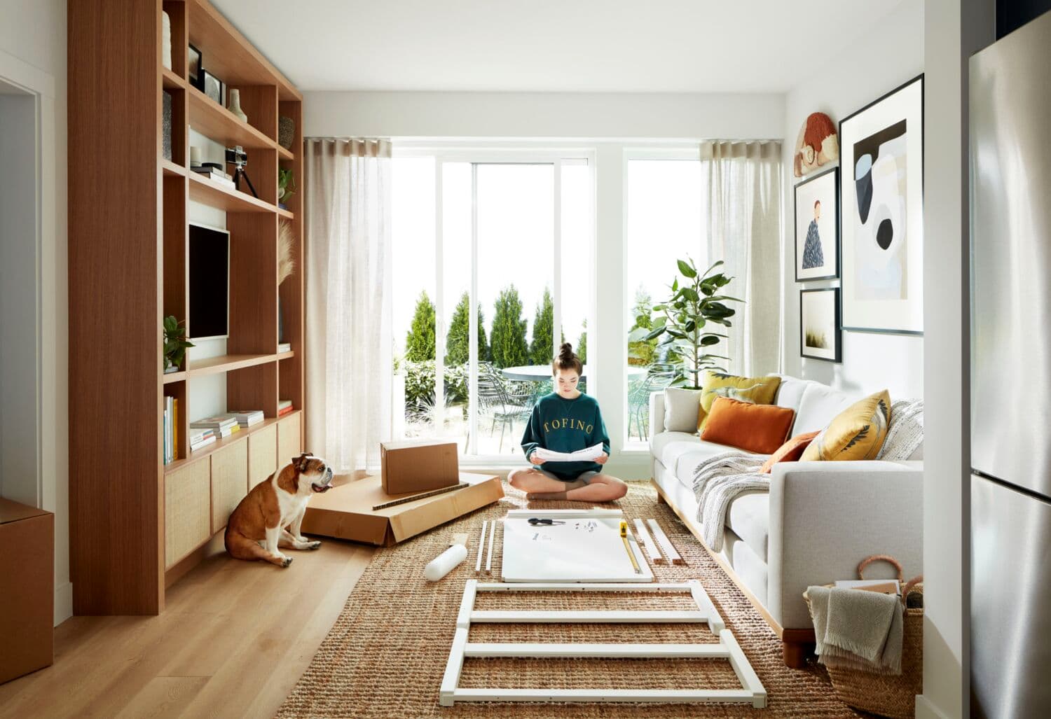 Moody Yards – A living room with a wood media wall, a light sofa, and a woman assembling furniture on a jute rug while her dog watches, with a patio visible through the window.
