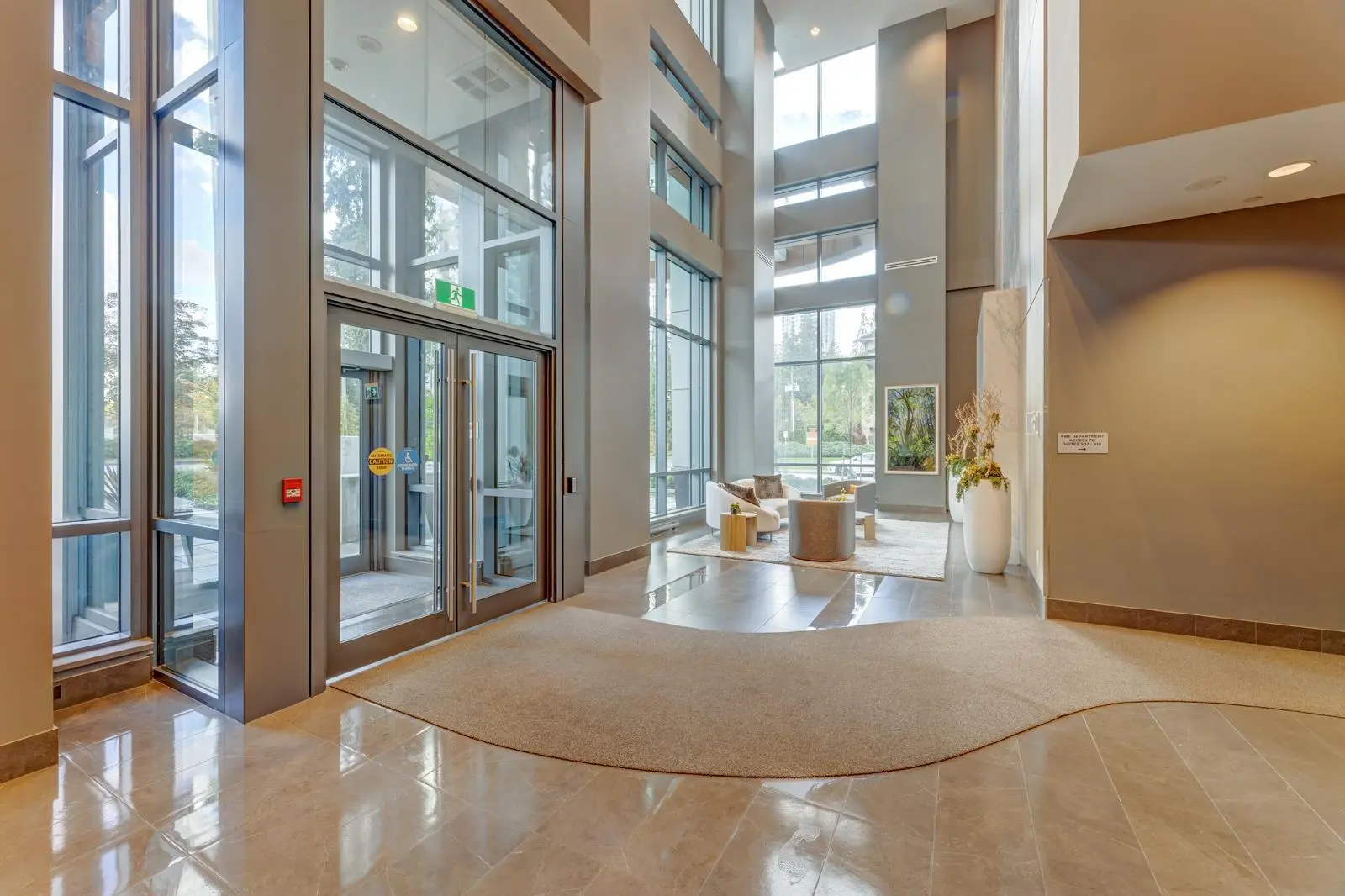 Sophora at the Park – Double-height building lobby entrance with glass doors, a curved area rug, and a seating area with armchairs visible beyond.