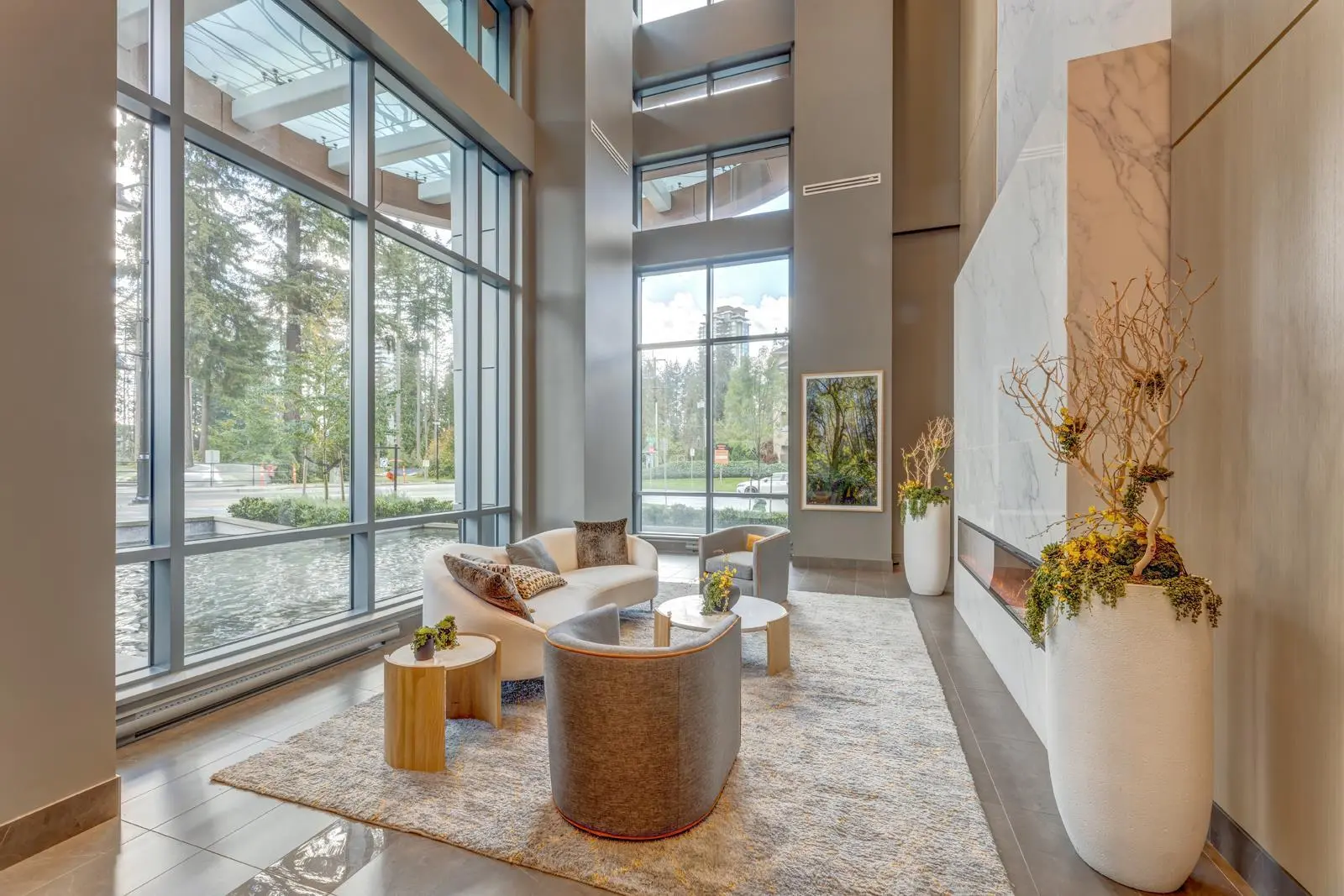 Sophora at the Park – Lobby lounge area with a curved sofa and armchairs around low tables, a marble fireplace wall, tall potted branches, and floor-to-ceiling windows facing a courtyard pond and street.