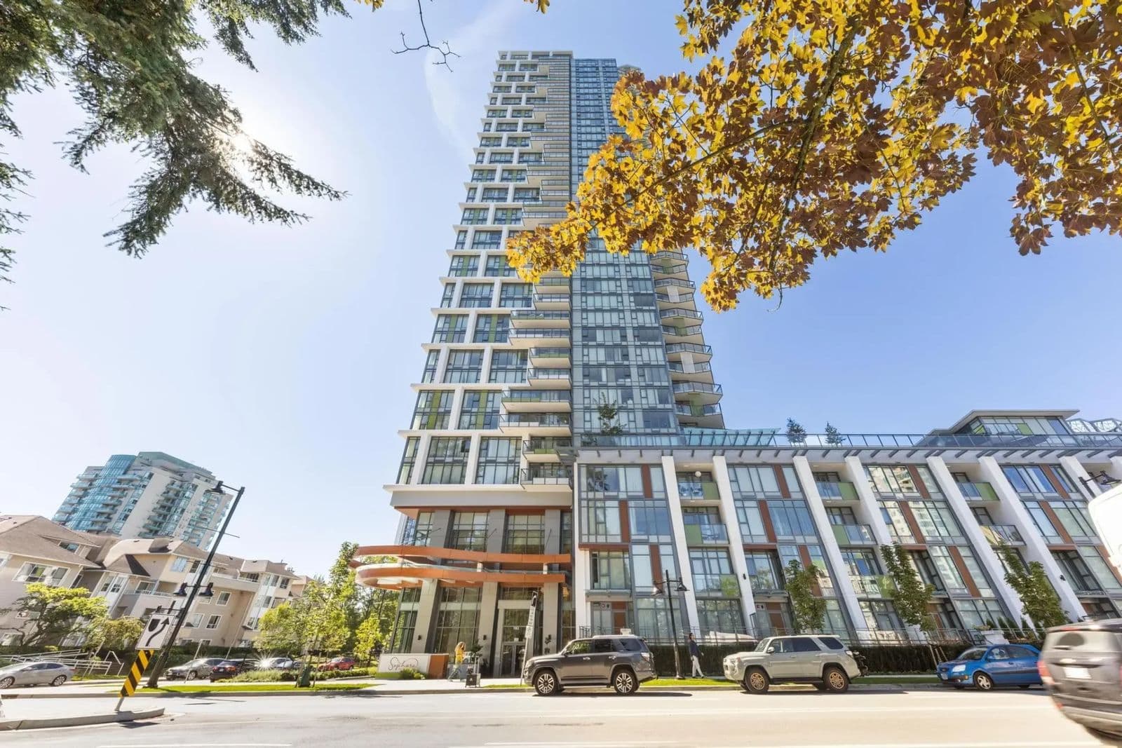 Sophora at the Park – Low-angle street view of the tower in autumn, with yellow foliage in the foreground, parked SUVs, and the building's entrance signage visible at street level.