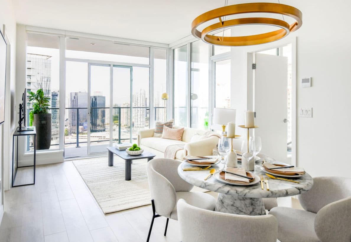 Riviera – Staged open-concept living and dining room in whites and neutrals with a marble dining table, brass ring chandelier, and floor-to-ceiling windows overlooking the Metrotown skyline.
