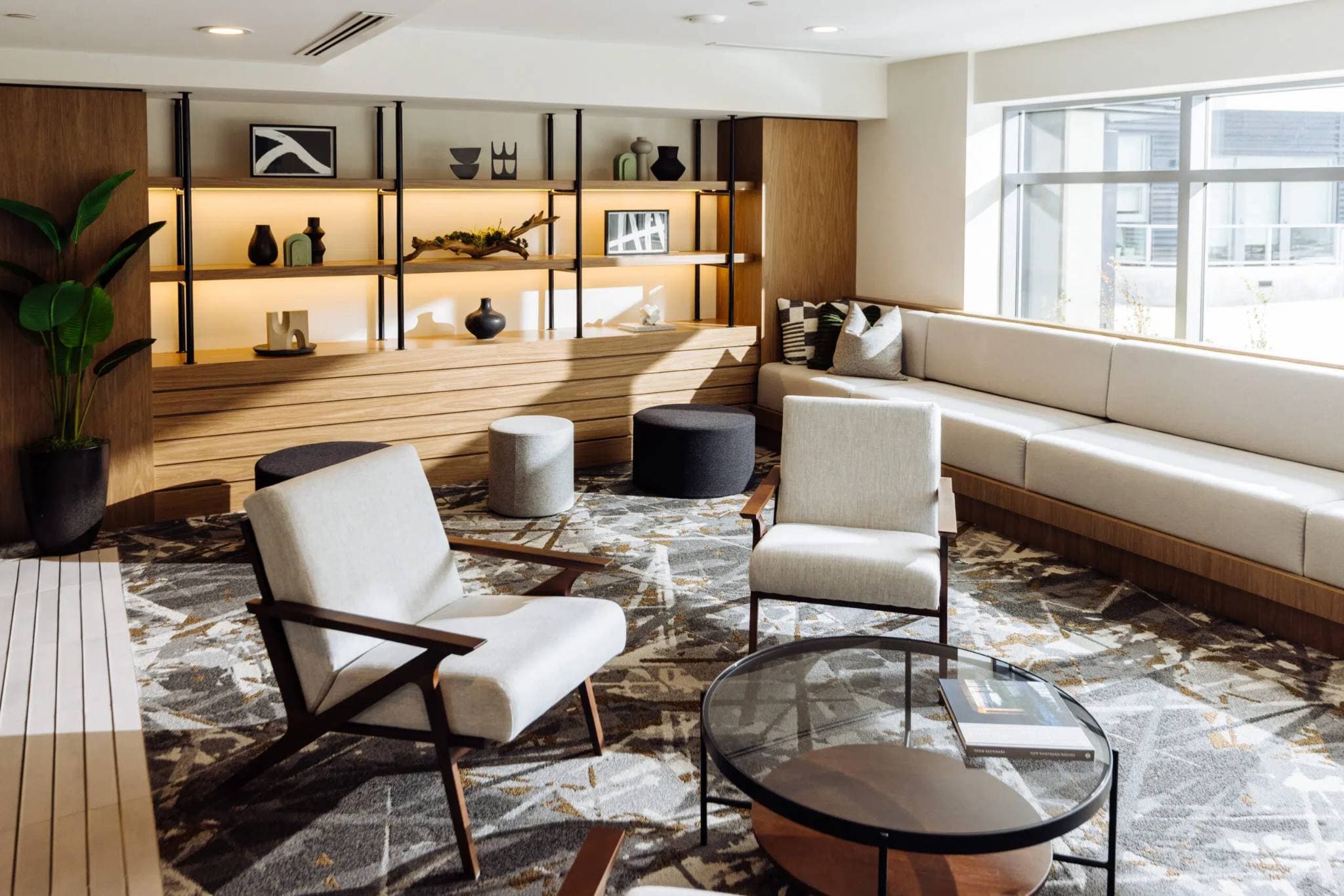 Paradigm – A modern residential lounge area featuring mid-century armchairs, a glass coffee table, and a long built-in sofa beside a large window. Warm LED backlighting highlights custom wooden shelving adorned with minimalist decor, creating a cozy and stylish atmosphere in bright daylight.