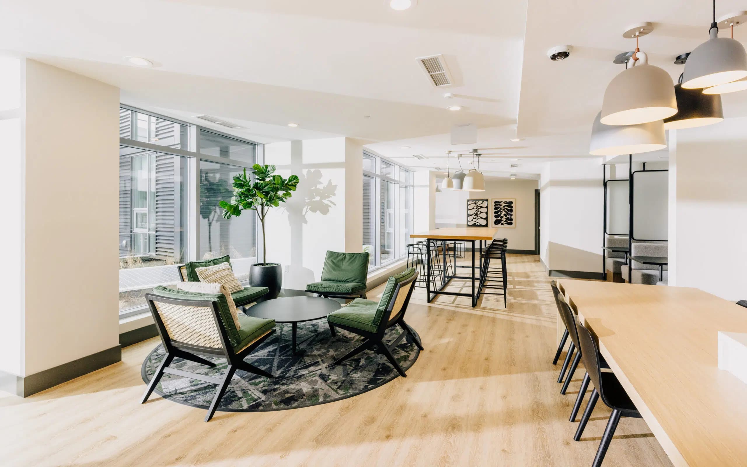 Paradigm – A bright, sunlit modern co-working lounge featuring light wood flooring, a seating area with green velvet armchairs, and long communal work tables under stylish pendant lighting. Floor-to-ceiling windows line the wall, casting soft daylight across the open-concept shared space.