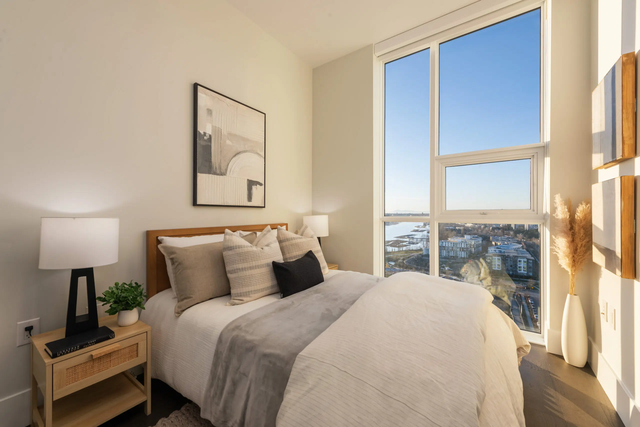 Paradigm – A modern bedroom setup featuring a wooden bed frame dressed in neutral layered linens, flanked by light-wood nightstands with modern table lamps. Floor-to-ceiling windows flood the bright space with natural light and offer expansive views of a river, city buildings, and clear blue sky.