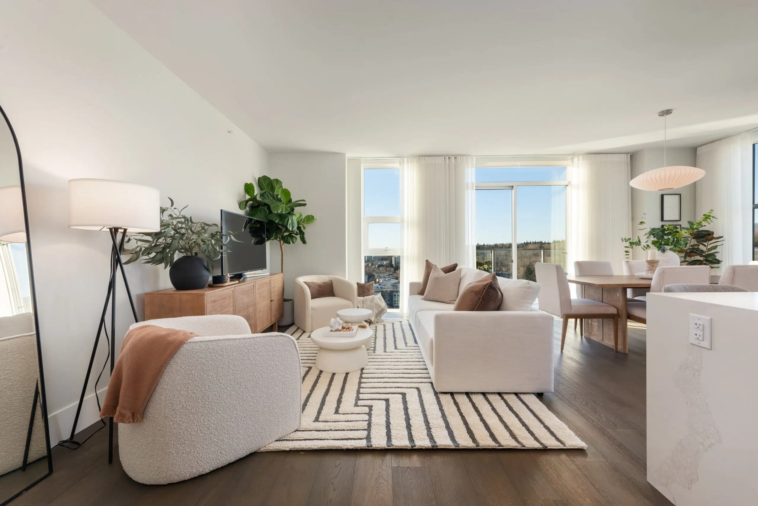 Paradigm – A bright, open-concept living and dining area featuring dark hardwood floors, a white textured sofa, bouclé accent chairs, and a geometric patterned area rug. Floor-to-ceiling windows with sheer white drapes let in natural daylight and offer views of surrounding trees and a distant skyline.