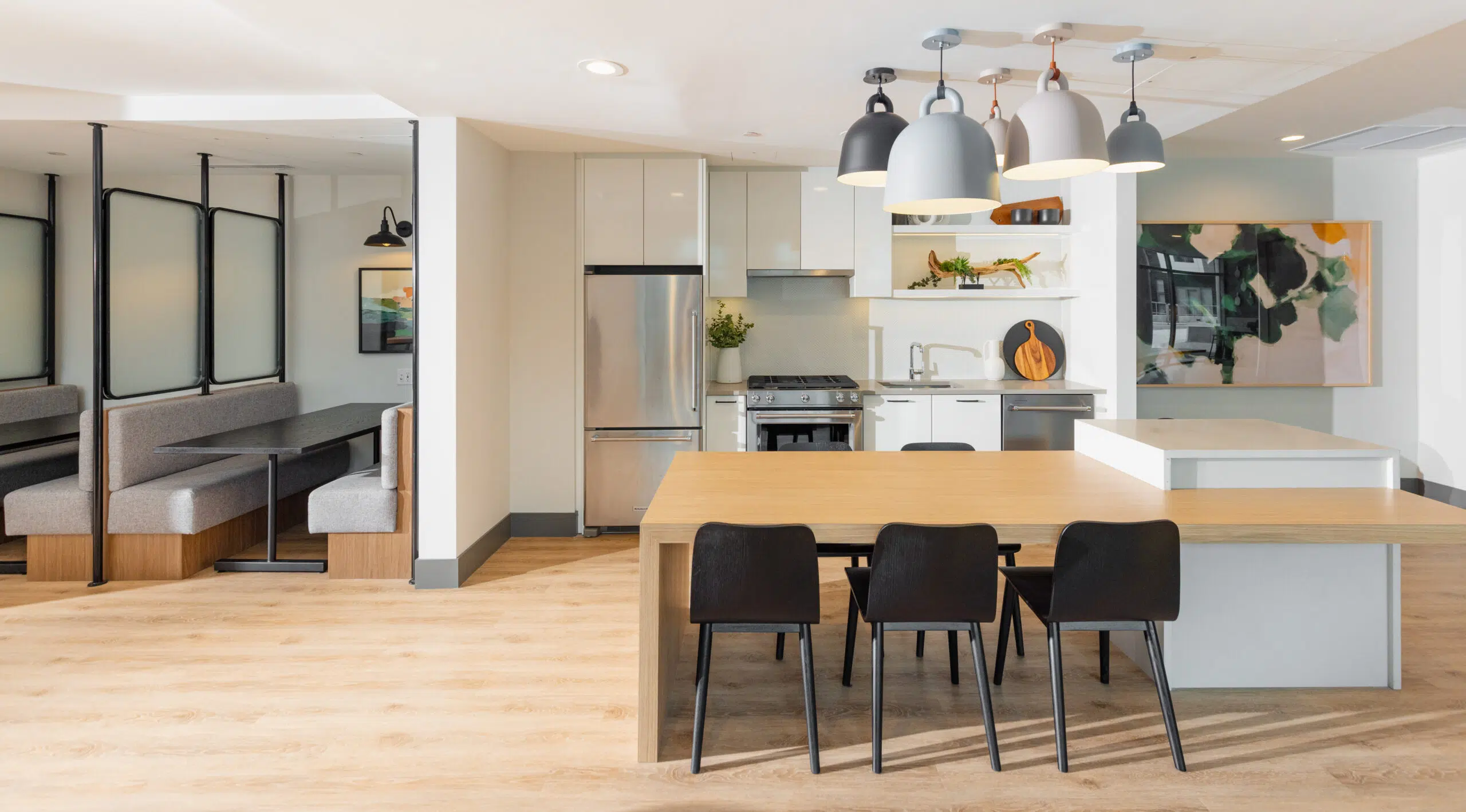 Paradigm – A modern communal kitchen and dining lounge featuring a light wood dining table with black chairs, hanging dome pendant lights, stainless steel appliances, and adjacent private seating booths enclosed by frosted glass partitions.