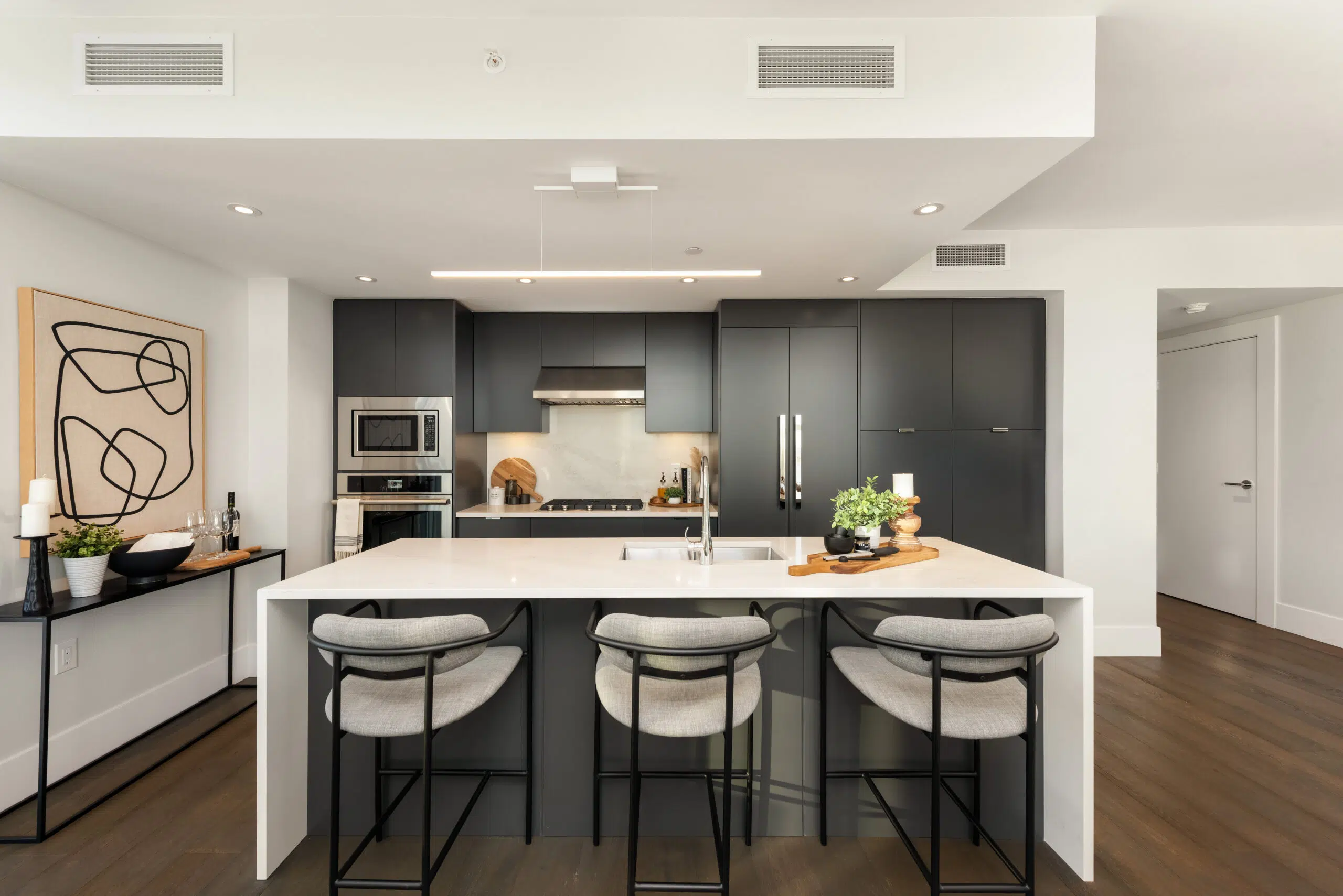 Paradigm – A modern open-concept kitchen features a large white waterfall island with three grey upholstered bar stools and matte black flat-panel cabinetry. Built-in stainless steel appliances, dark wood flooring, and clean minimalist decor complete the sophisticated residential interior.