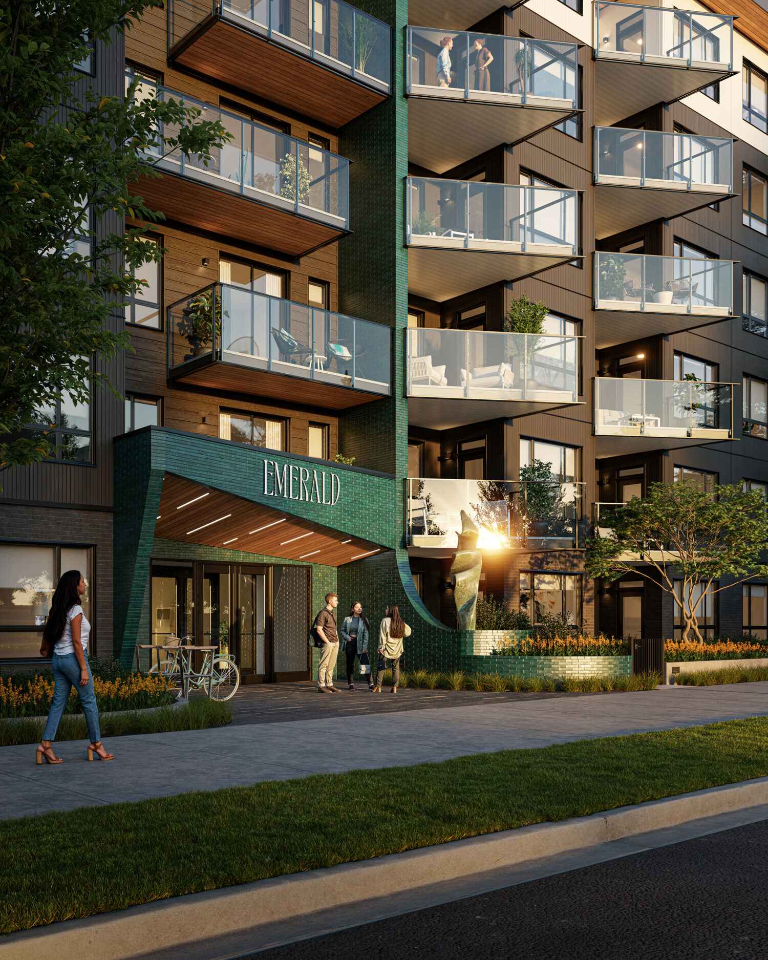 Emerald – A ground-level view of a modern multi-story residential building featuring dark siding, wood-toned soffits, and a distinctive entrance canopy wrapped in glossy teal tiles labeled EMERALD. Pedestrians walk along the paved sidewalk and landscaped garden beds under warm, late-afternoon sunlight.