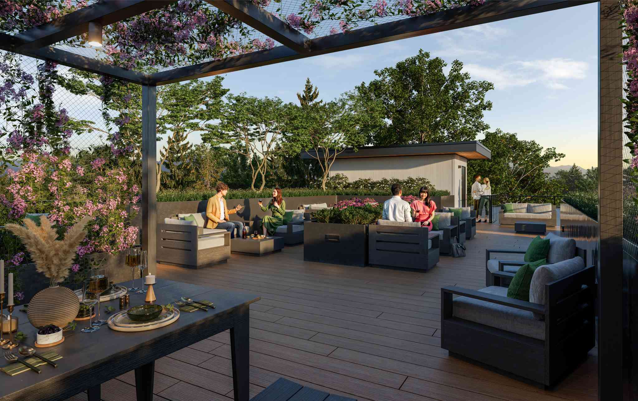 Emerald – A wood-decked rooftop terrace features multiple lounge seating areas surrounded by lush planters and a pergola draped with purple flowering vines. People gather in casual groups enjoying drinks under a bright, clear sky bordered by lush green treetops.