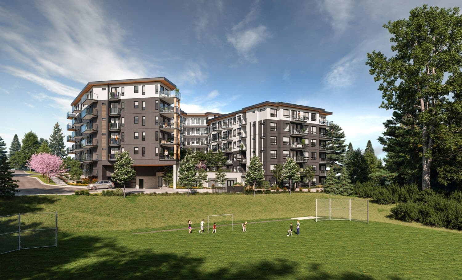 Emerald – A modern mid-rise residential building with charcoal grey and white siding, glass balconies, and wood accents stands under a bright, partly cloudy sky. In the foreground, a grassy sports field features children playing soccer near mini nets, bordered by lush green trees and a paved street.