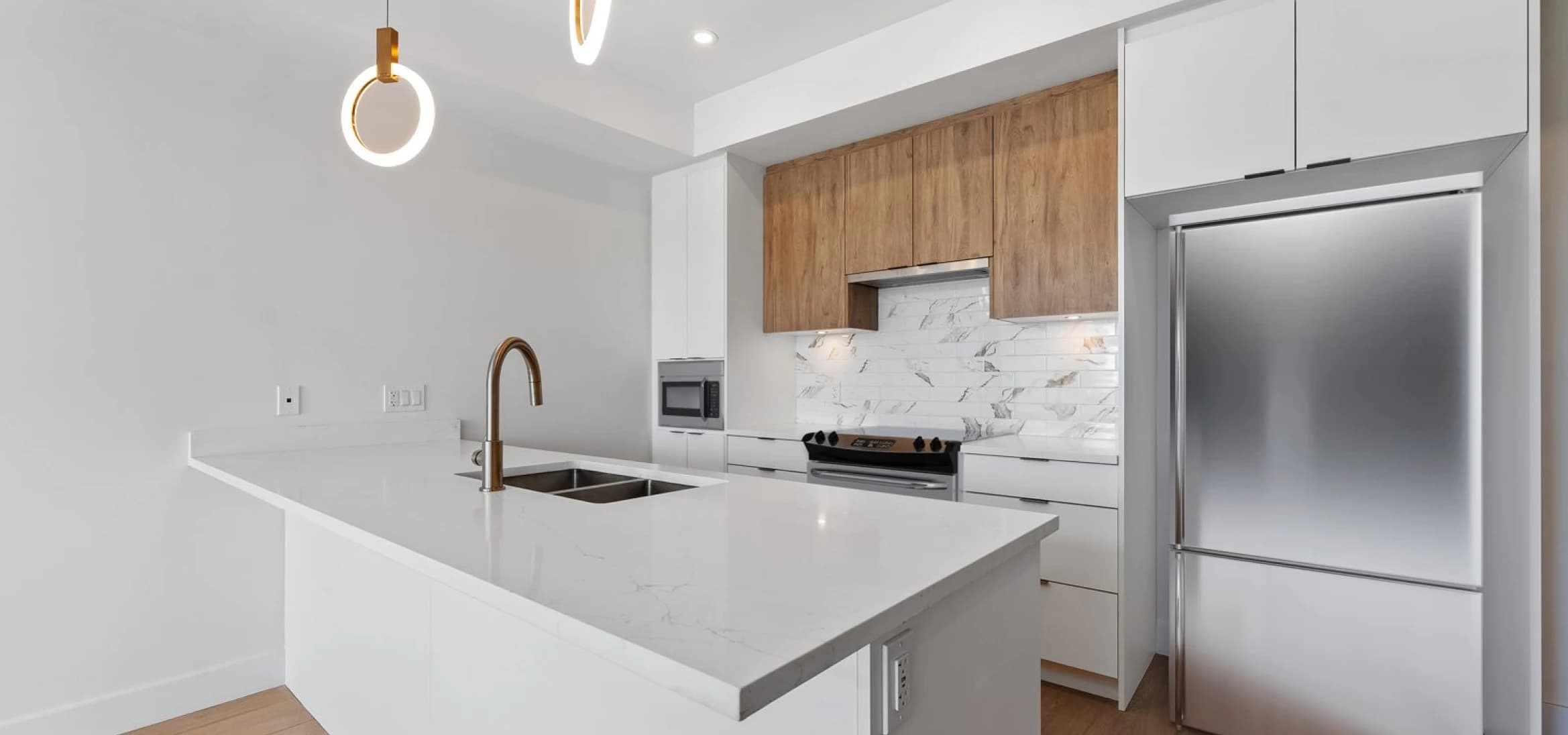 Kingsgate – A modern kitchen featuring a large white quartz island with an undermount double sink and gold gooseneck faucet beneath halo pendant lights. In the background, two-tone cabinetry combines warm wood upper cabinets with sleek white lowers, complemented by a marble tile backsplash and stainless steel appliances.