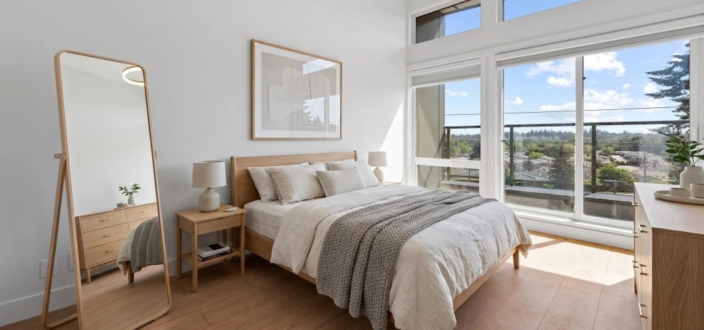 Kingsgate – A sunlit contemporary bedroom featuring a light wood bed dressed in neutral white and grey linens, flanked by matching nightstands and a full-length standing mirror. Floor-to-ceiling sliding glass doors open onto a balcony overlooking treetops and a clear blue sky.