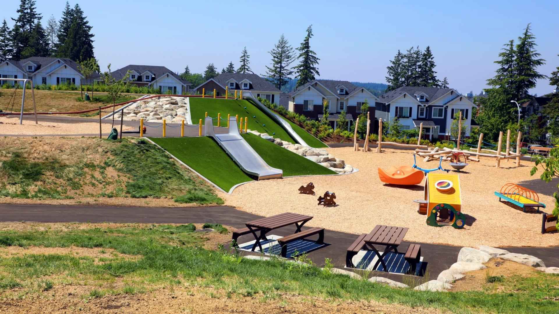 Kingsgate – A spacious outdoor community playground and park featuring embankment slides built into turf-covered hills, rock climbing areas, modern play structures, and picnic tables under bright daylight. In the background, newly built residential townhomes and single-family houses are framed by tall evergreen trees.