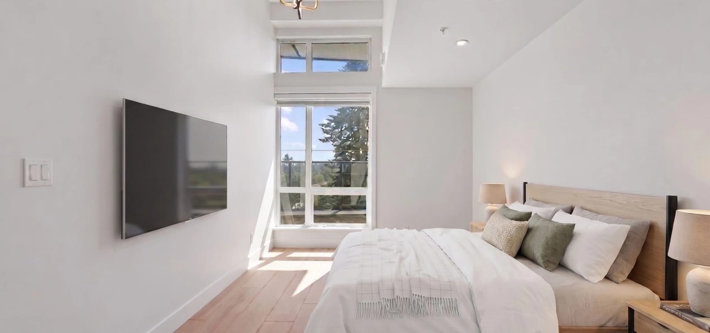 Kingsgate – A bright, minimalist bedroom features a neatly made bed with light wood accents, white bedding, and muted green pillows, set against crisp white walls and light hardwood floors. Sunlight pours in through a tall, transom-topped window at the far end of the room, looking out onto lush green trees and a balcony.