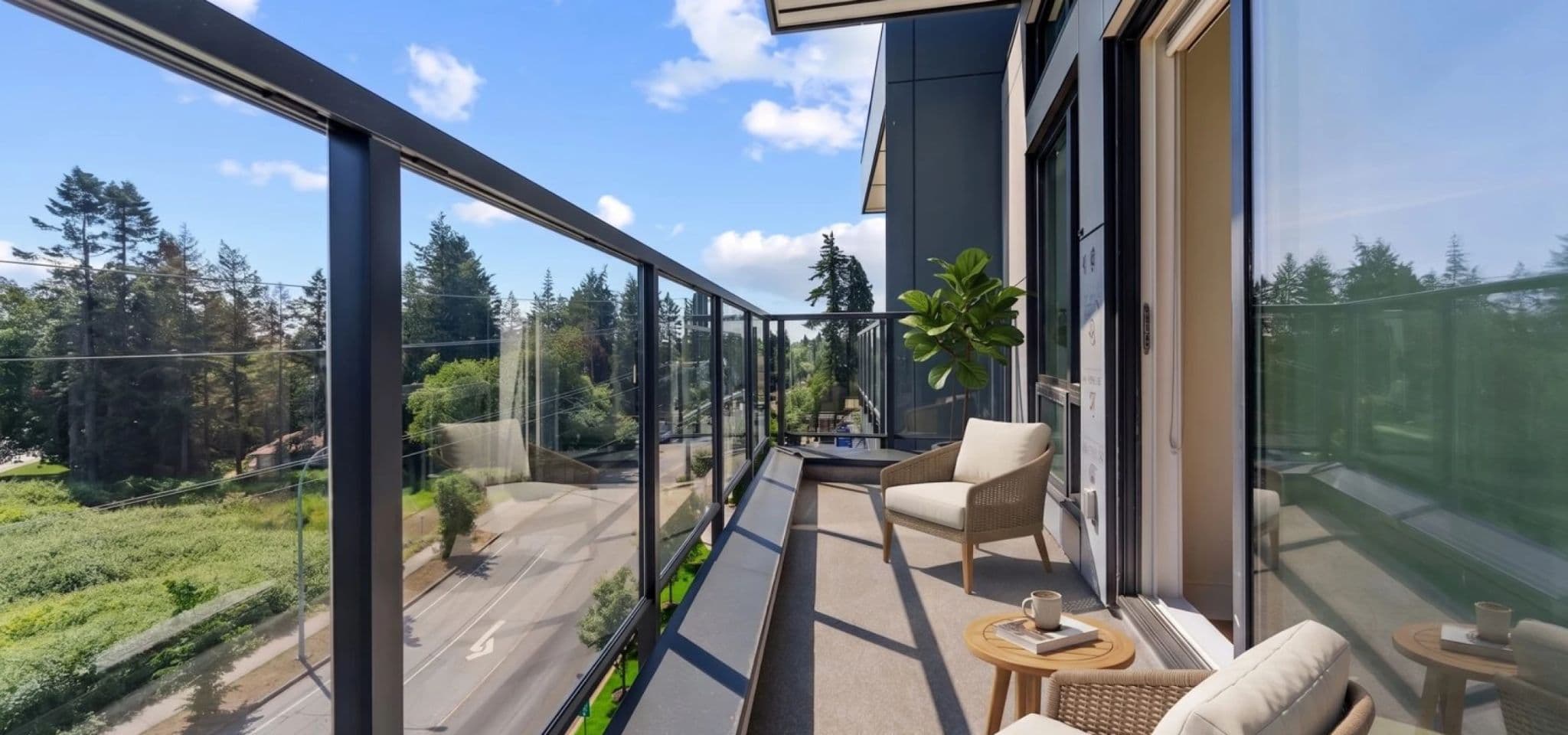 Kingsgate – A modern glass-railed private balcony furnished with woven armchairs, a small side table, and a potted plant, offering an open view over a quiet tree-lined street under a bright, partly cloudy sky. The sunlit outdoor terrace overlooks dense evergreen foliage and surrounding residential nature.