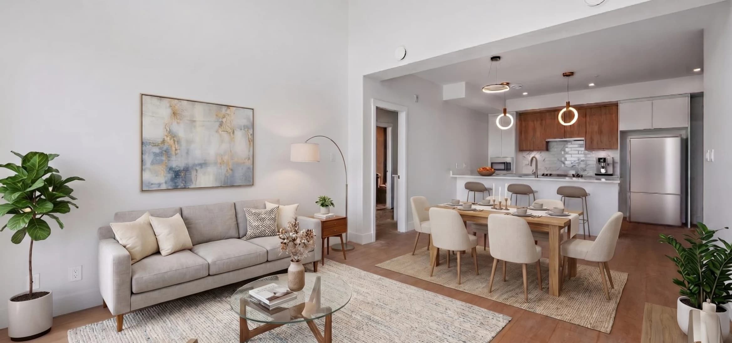 Kingsgate – A bright, open-concept living space features a light gray sofa set over a textured rug, alongside a wooden dining table paired with cream upholstered chairs. In the background, an open kitchen displays warm wood cabinetry, a breakfast island with barstools, and modern ring pendant lights.