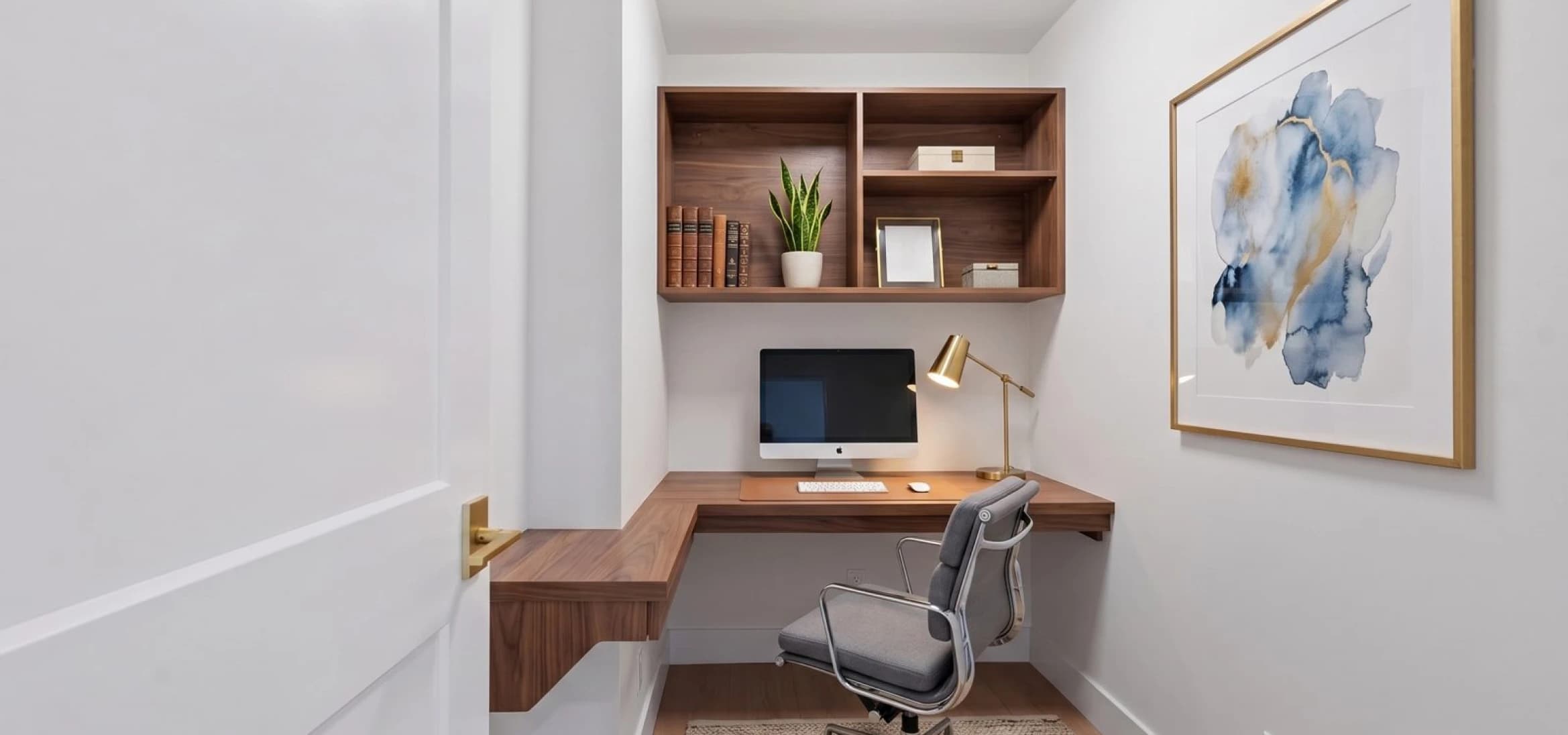 Kingsgate – A compact, modern home office nook featuring a built-in floating walnut desk and matching upper shelving unit set between crisp white walls. The workspace is styled with a desktop computer, a brass task lamp, a grey swivel chair, and a framed abstract blue-and-gold art piece.