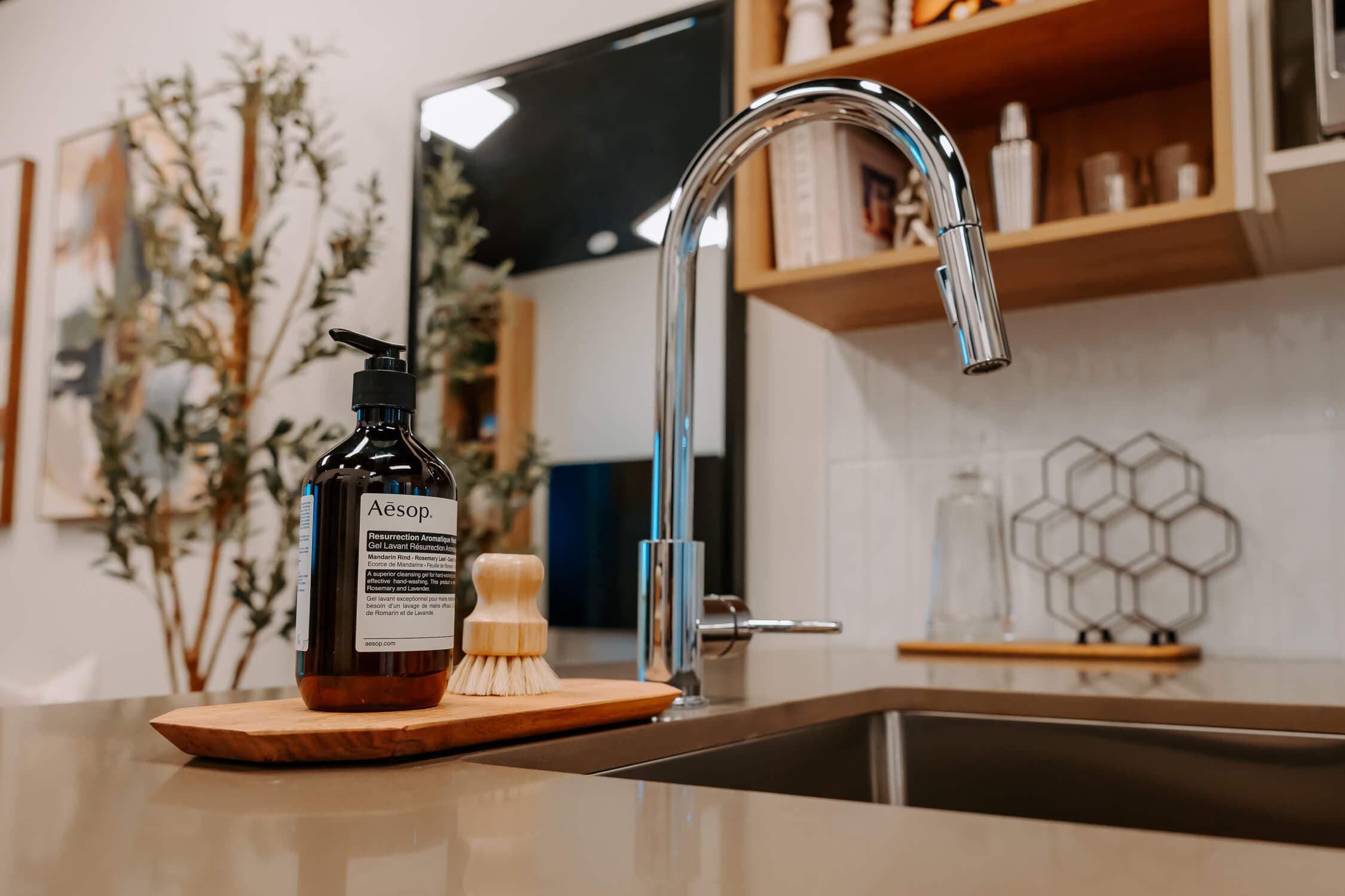 Stowa – A close-up view of a modern kitchen counter featuring an arched chrome faucet over an undermount sink. A bottle of Aesop hand soap sits on a small wooden dish next to a wooden dish brush, creating a warm, styled domestic aesthetic.