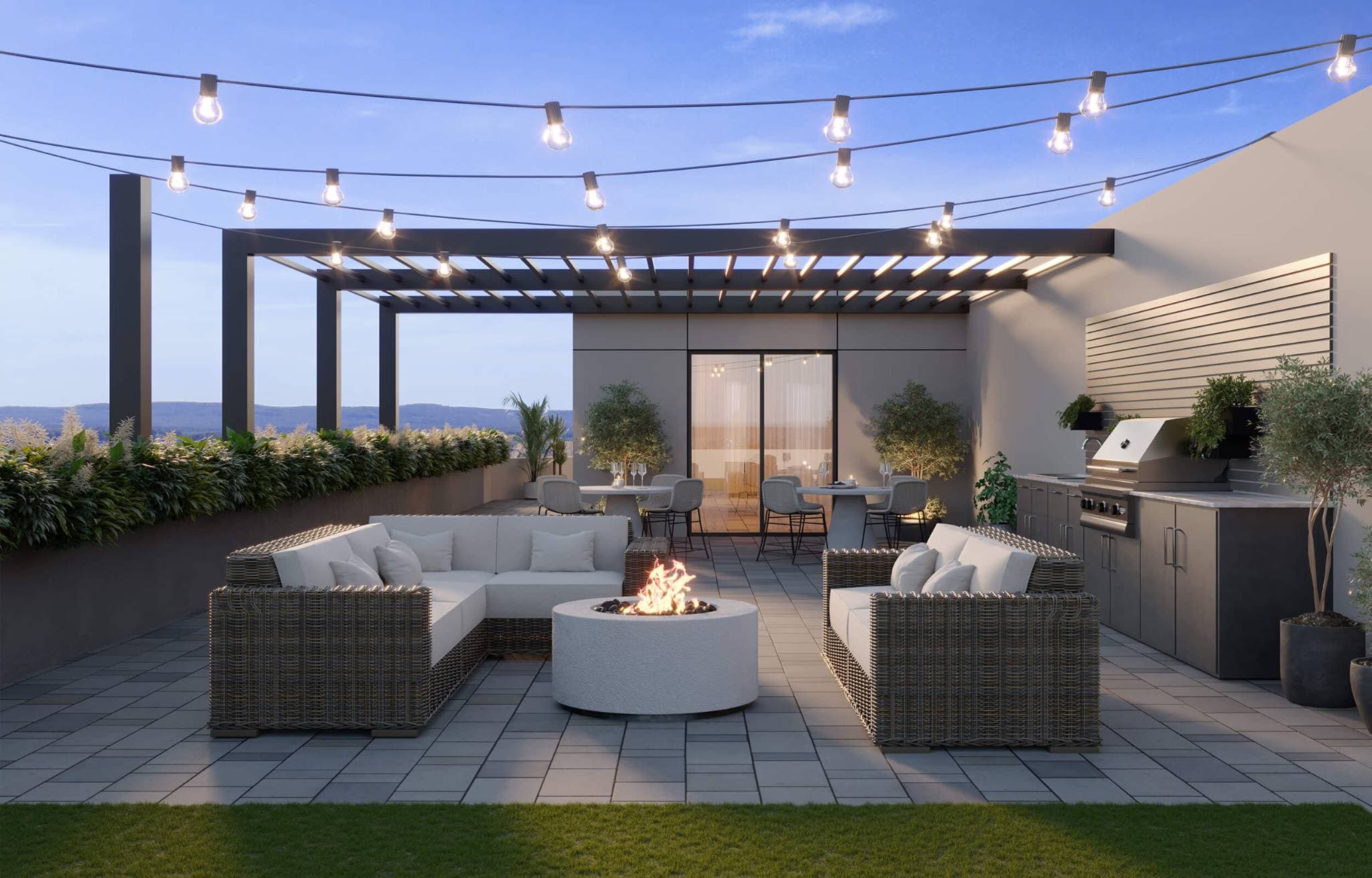 Stowa – A twilight rooftop terrace features woven outdoor sofas surrounding a round white fire pit, string lights overhead, and a covered pergola over dining tables. An outdoor kitchen with a stainless steel grill sits along a side wall, while landscaped planters frame views of distant mountains under a soft blue sky.