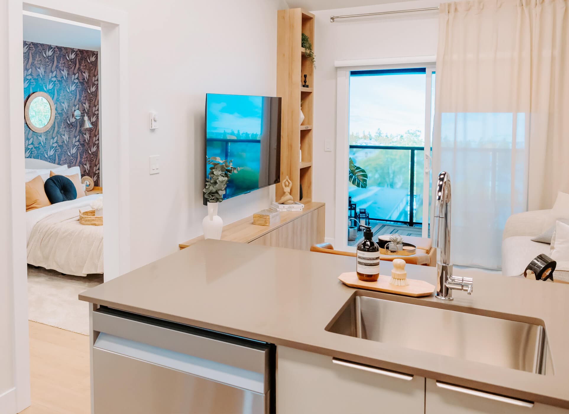 Stowa – A modern open-concept condo interior featuring a kitchen island with an undermount sink in the foreground leading into a bright living room with a wall-mounted TV, light wood console, and sliding glass doors to a balcony. To the left, an open doorway reveals a cozy bedroom styled with patterned wallpaper, accent pillows, and warm neutral tones.