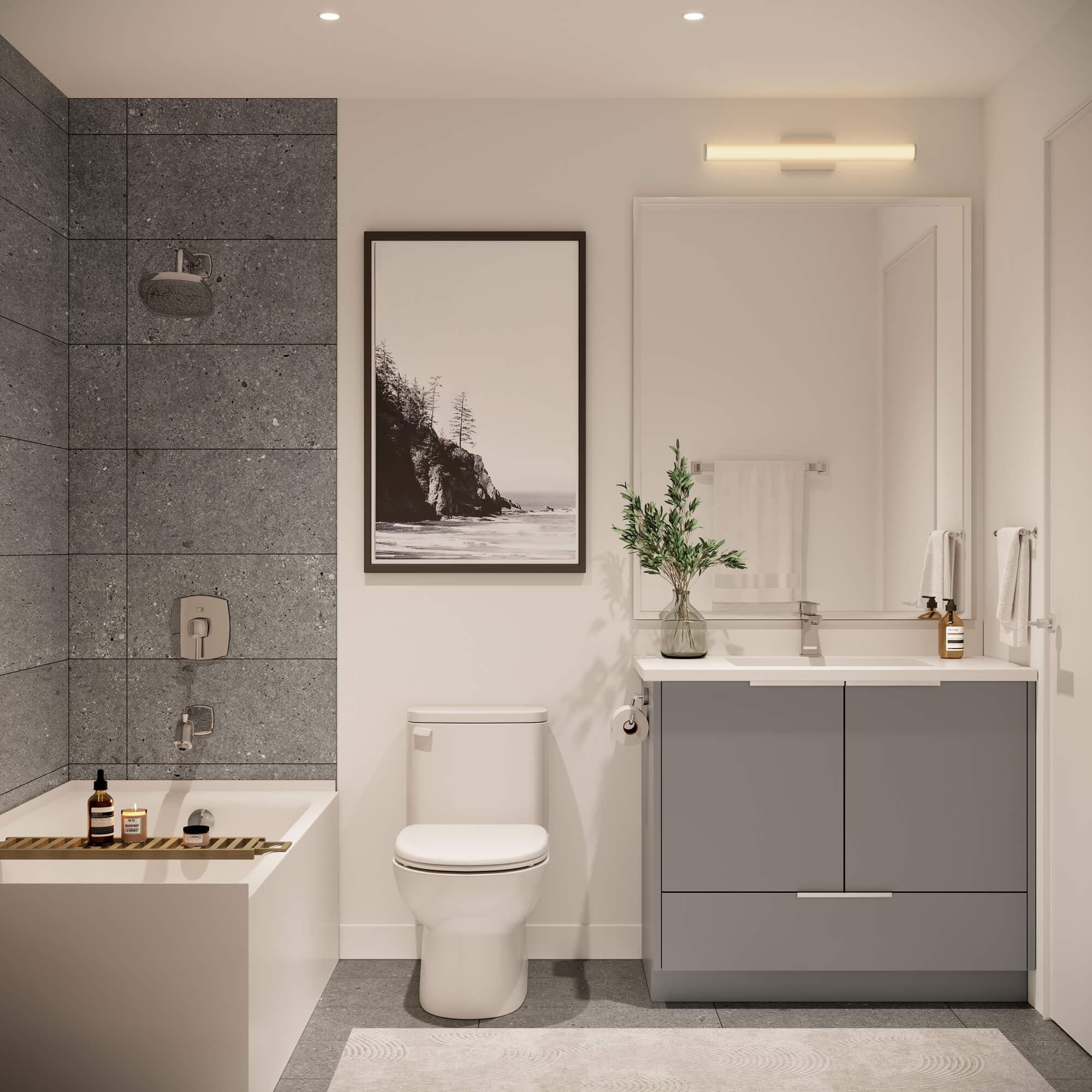 Stowa – A modern bathroom featuring a grey floating vanity with a white countertop, a white toilet, and a bathtub surrounded by dark grey terrazzo tiles. Warm lighting, a framed black-and-white landscape print, and minimalist accessories create a bright and serene atmosphere.