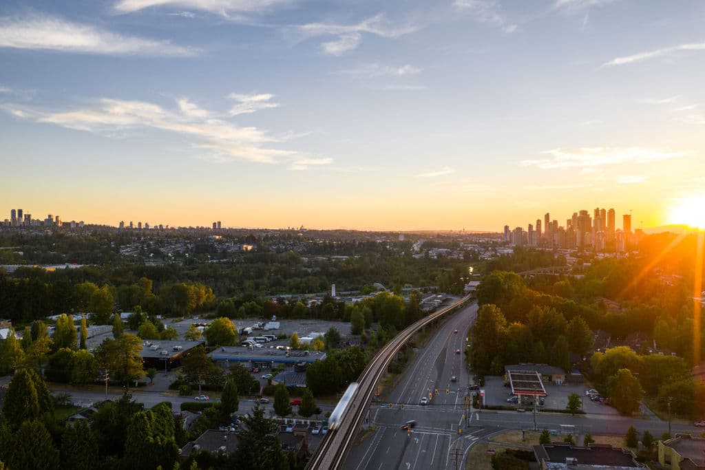 Blake Village – An elevated aerial view captures a transit rail line and roadway cutting through lush green suburban neighborhoods toward distant high-rise skylines during a vibrant golden hour sunset.