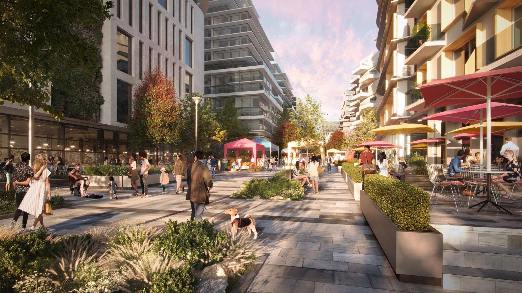 Blake Village – A ground-level render of a lively pedestrian plaza lined with modern multi-story buildings, outdoor cafe dining under vibrant umbrellas, and landscaped planter boxes. People and pets stroll along the stone walkway past pop-up market stalls in warm, late-afternoon sunlight.