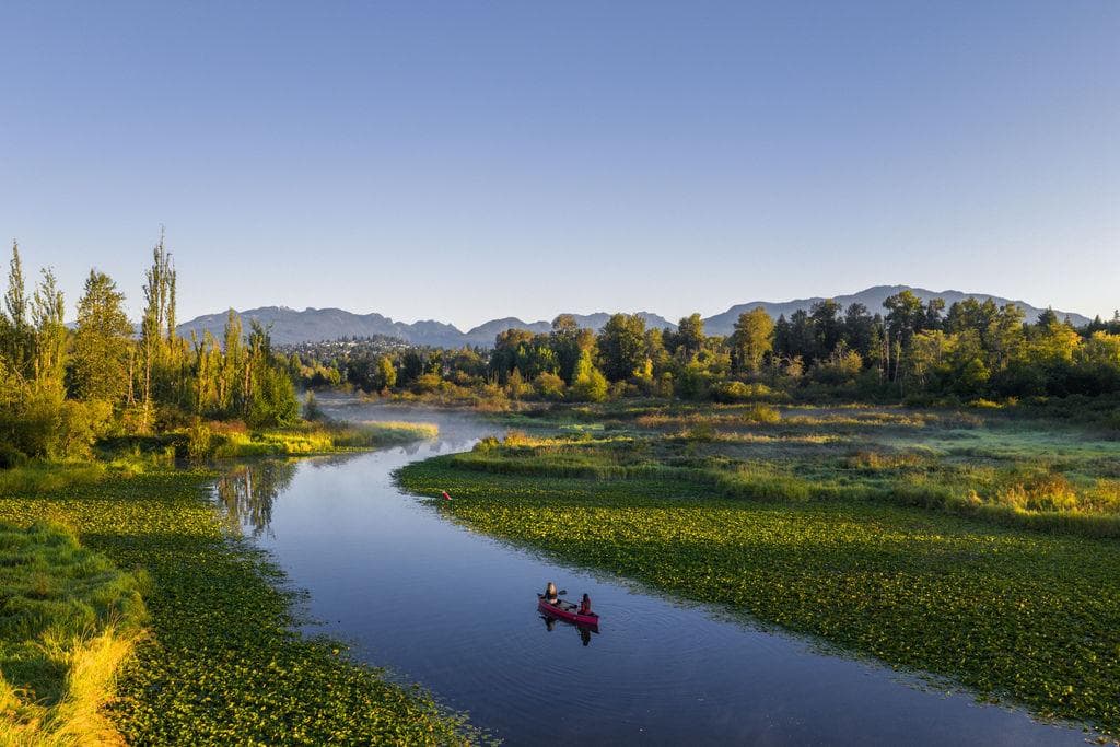 Blake Village – An elevated view of a canoe paddling down a winding, calm river bordered by wetlands and lush green trees. In the background, distant mountains sit under a clear blue sky in bright morning light.