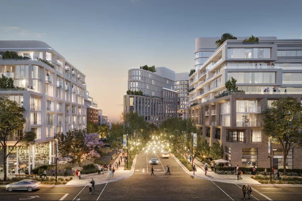 Blake Village – A dusk view looking down a central, tree-lined street bordered by modern mid-rise residential buildings with curved facades, ground-floor retail, and rooftop greenery. Warm string lights canopy over the roadway as pedestrians stroll along wide sidewalks.