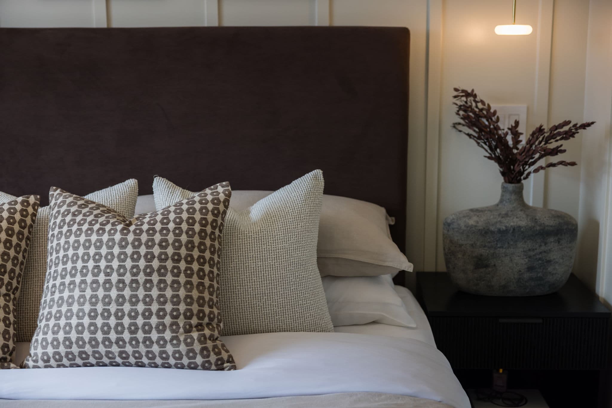 Charlton – Close-up of a dark upholstered headboard with patterned and textured pillows next to a nightstand holding a textured vase.