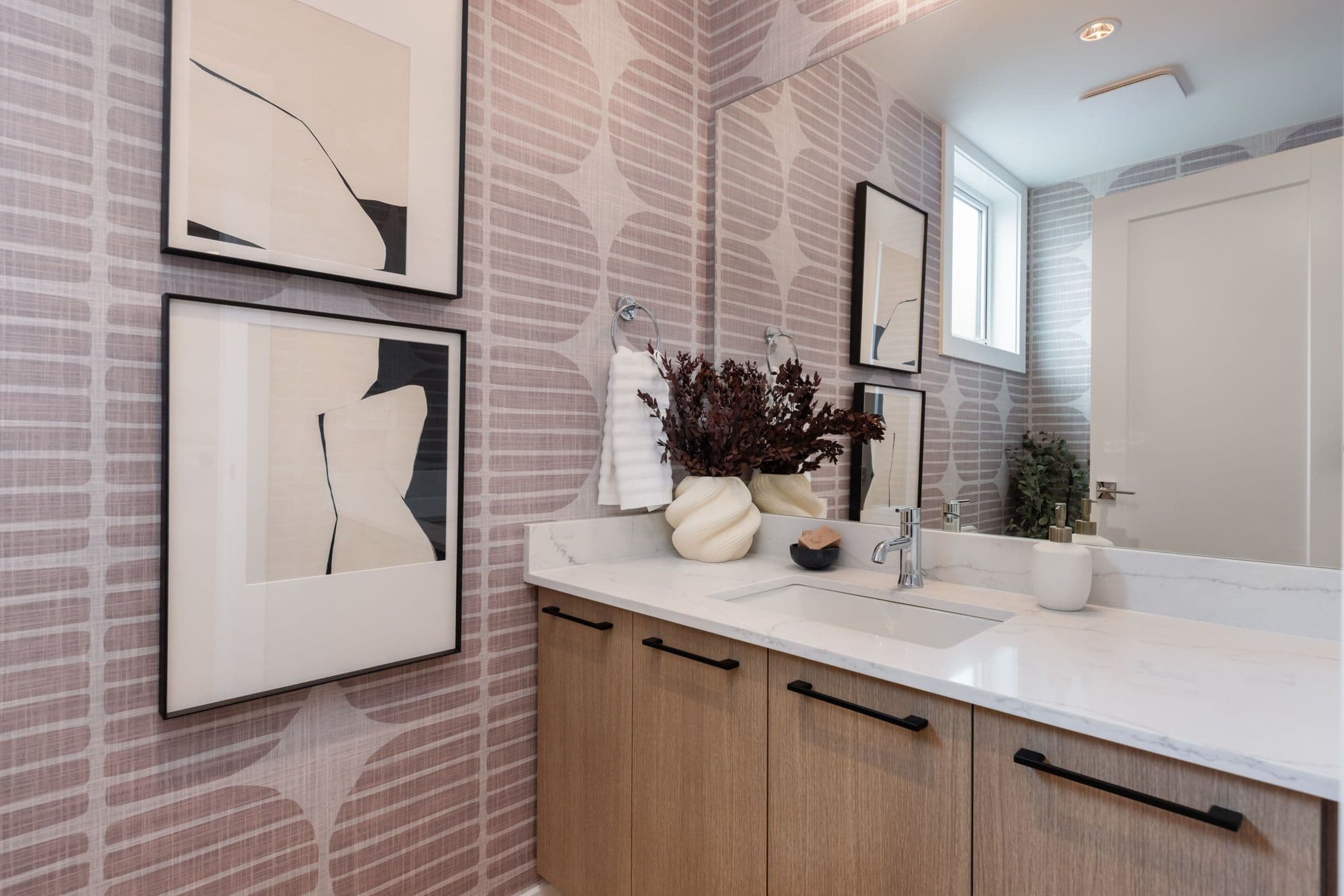Charlton – Bathroom with mauve geometric wallpaper, wood-tone vanity, abstract black-and-white framed art, and a vase of dried branches.