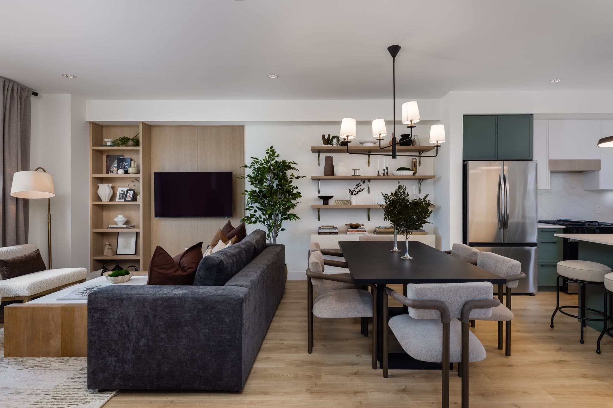 Charlton – Open-concept living and dining area with a grey sofa, dark dining table with upholstered chairs, and a view into a green-cabinet kitchen.