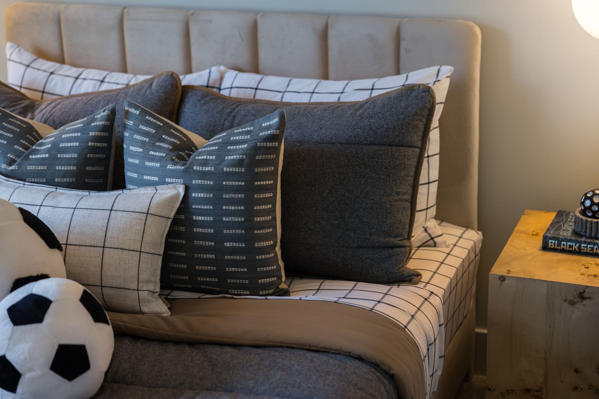 Charlton – Close-up of a bed styled with grey and windowpane-check pillows and a plush soccer ball, next to a wood nightstand with a book.