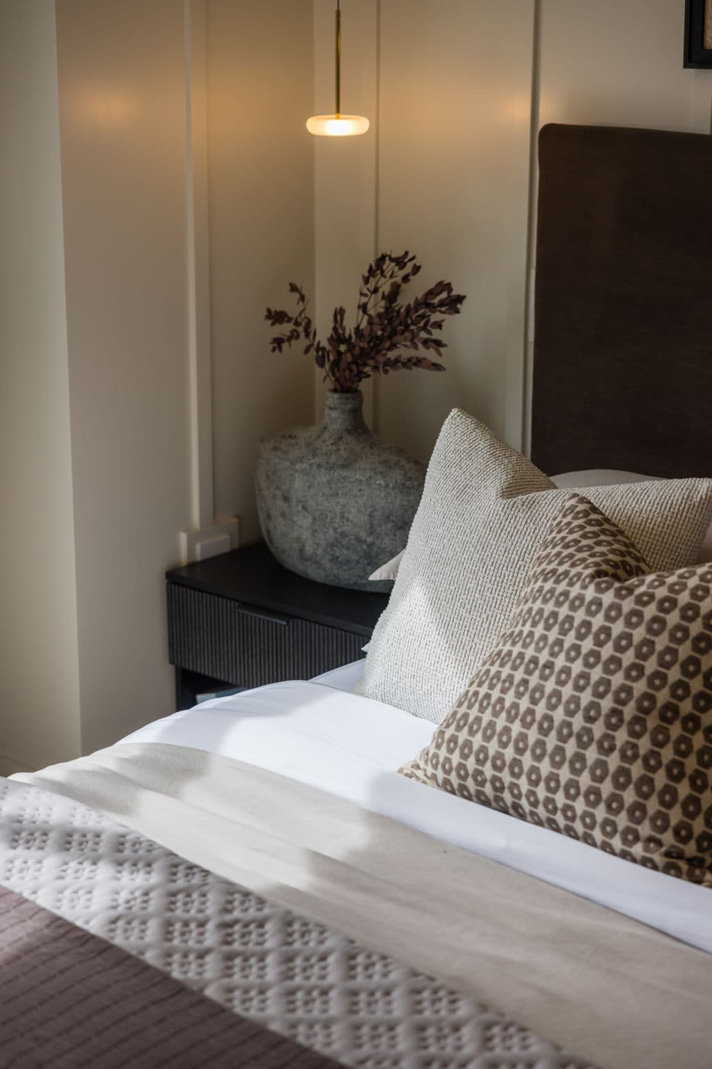 Charlton – Close-up of a dark upholstered headboard with textured and patterned pillows beside a black nightstand with a stone vase.
