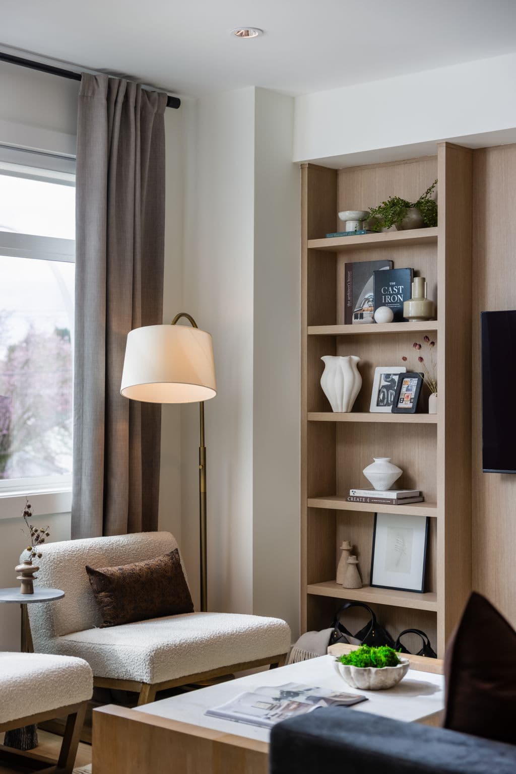 Charlton – Living room corner featuring a tall wood bookshelf with books and decor, a floor lamp, and a boucle armchair by the window.