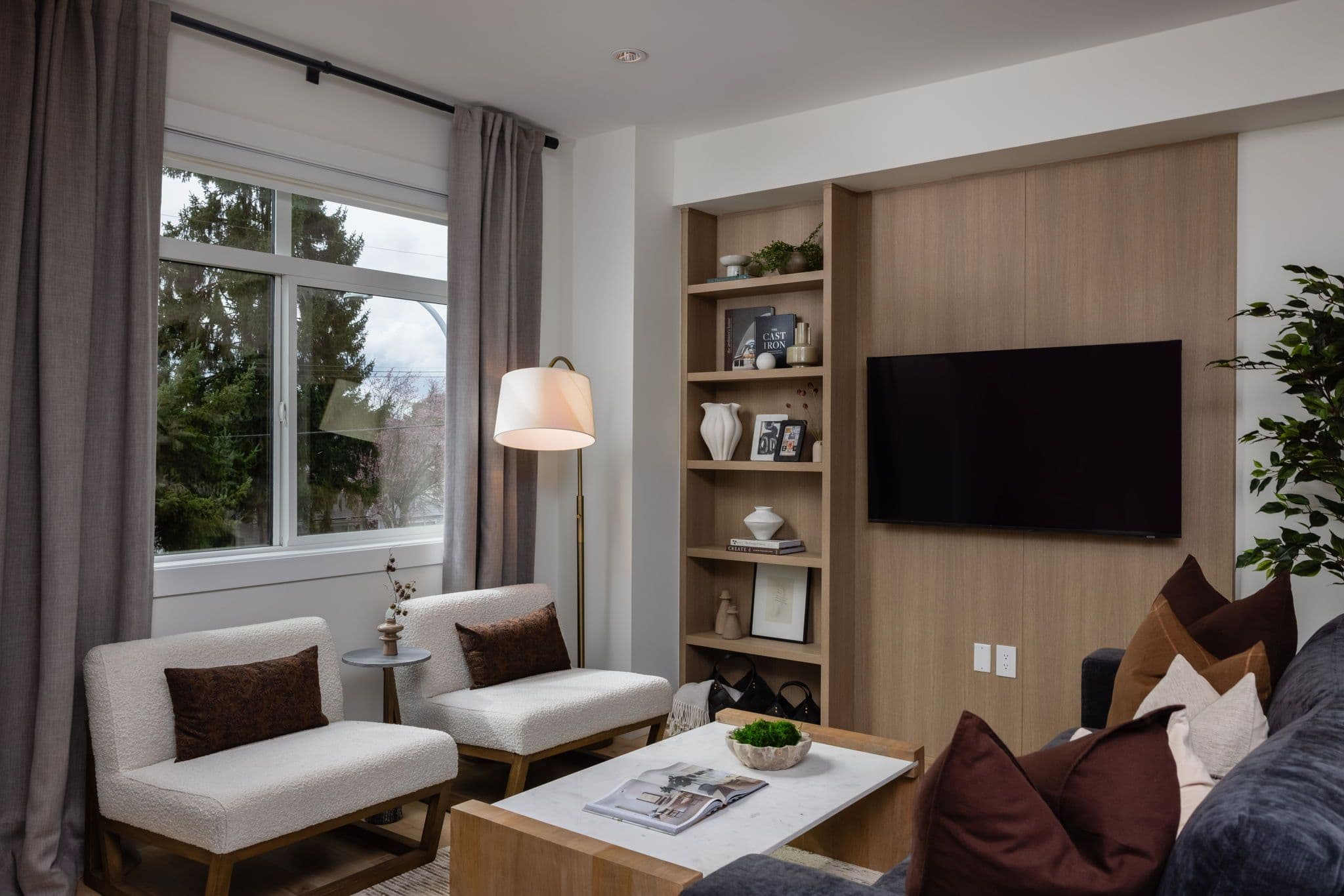 Charlton – Living room corner with a built-in wood bookshelf holding books and decor, a wall-mounted TV, and two boucle armchairs by a window.
